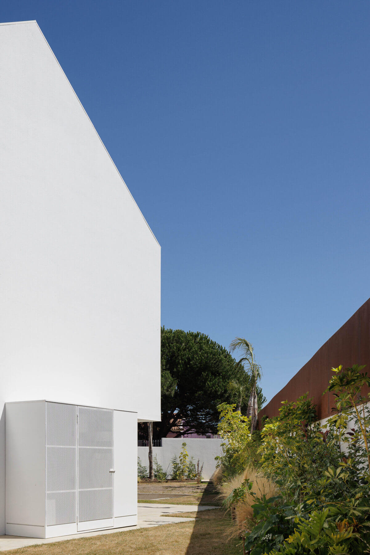 Jardim: Vista do jardim com relva, vegetação e anexo branco com porta em grelha metálica. — Casas BD arquitetura Esquissos Arquitectura em Sintra