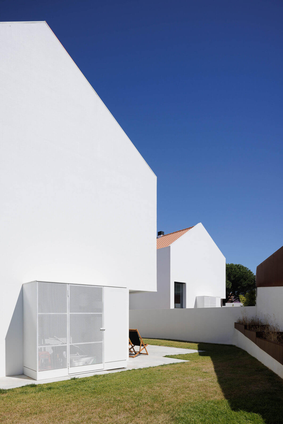 Jardim: Espaço exterior com relva, espreguiçadeira e anexo branco com porta em grelha metálica. — Casas BD arquitetura Esquissos Arquitectura em Sintra