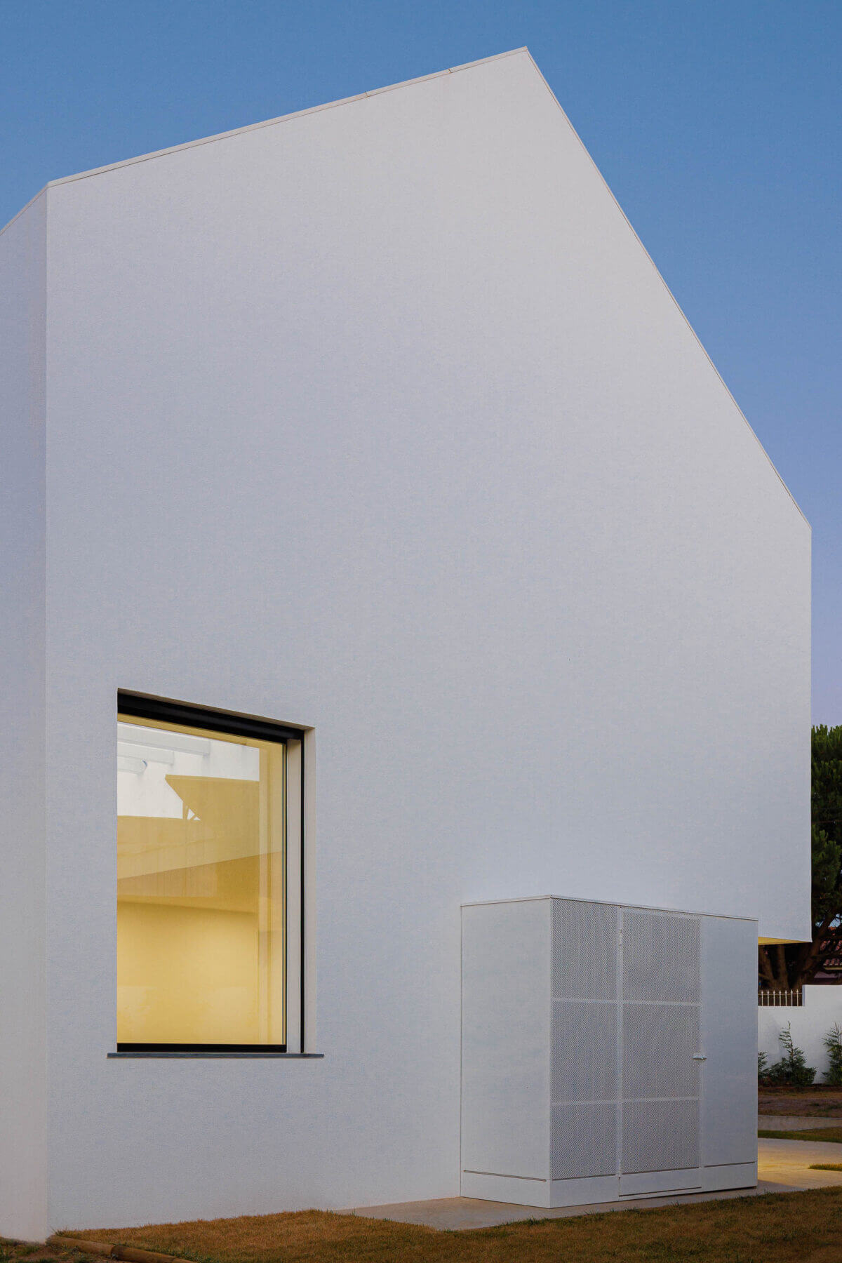Fachada: Moradia branca com janela iluminada e anexo com porta em grelha metálica ao entardecer. — Casas BD arquitetura Esquissos Arquitectura em Sintra