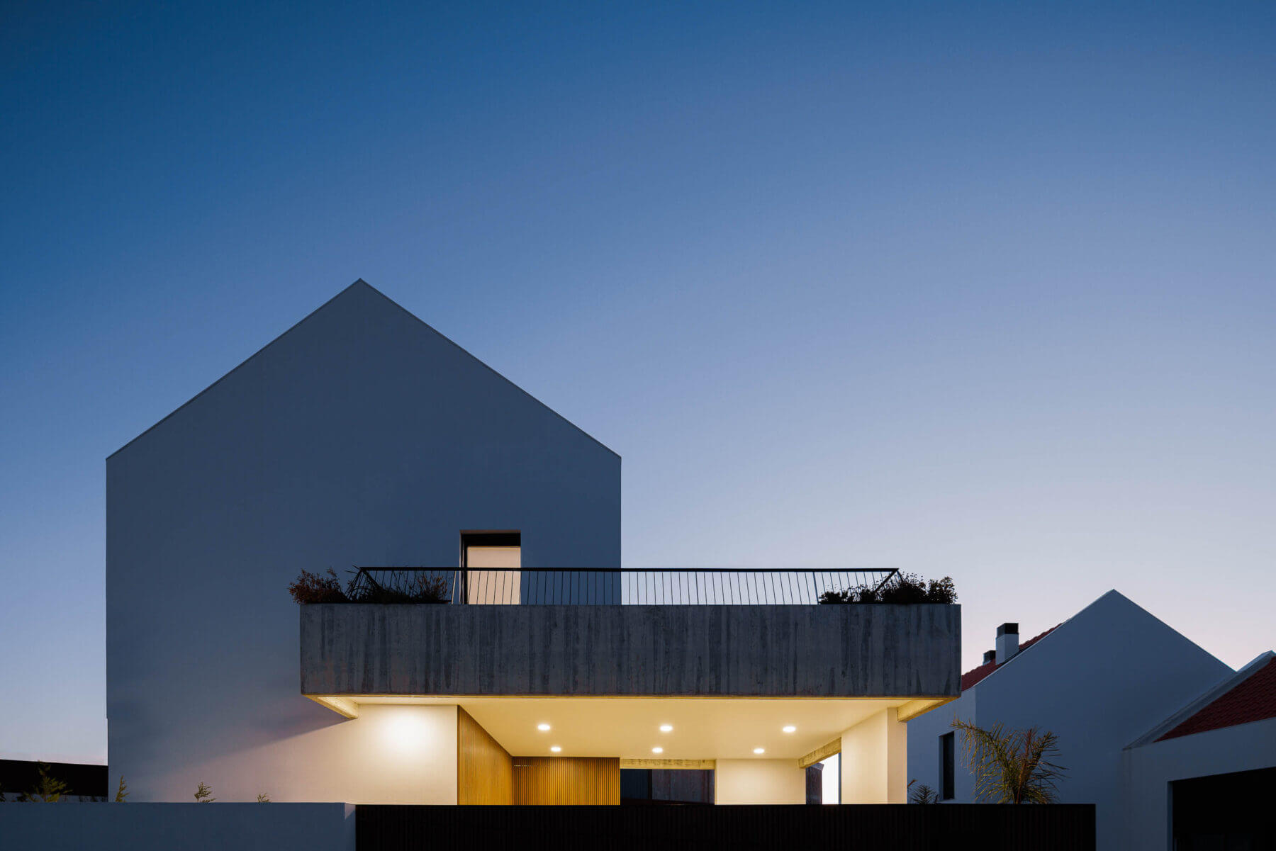 Fachada: Moradia branca com varanda em betão aparente iluminada ao anoitecer. — Casas BD arquitetura Esquissos Arquitectura em Sintra