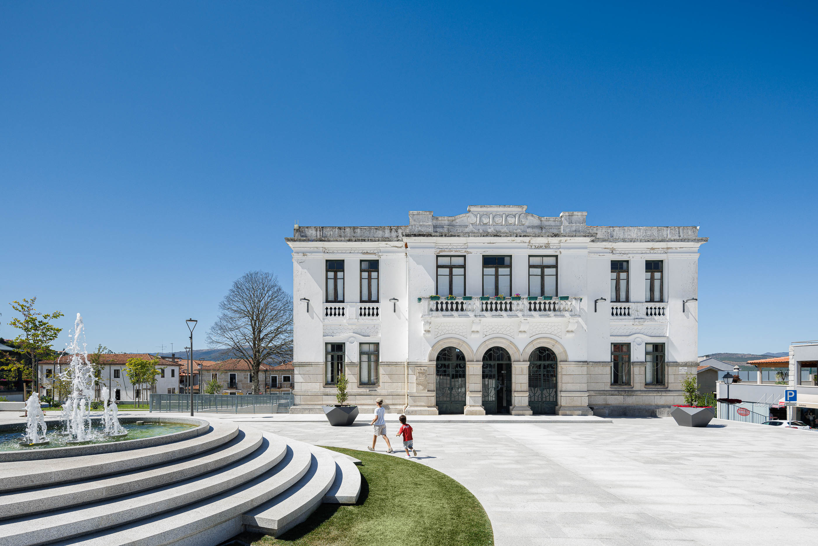 Praça: Edifício histórico branco com varanda, fonte escalonada, pavimento em granito e relvado. — Requalificação da Praça do Município de Ribeira de Pena arquitetura AXR - Arquitetura e Design em Vila Real