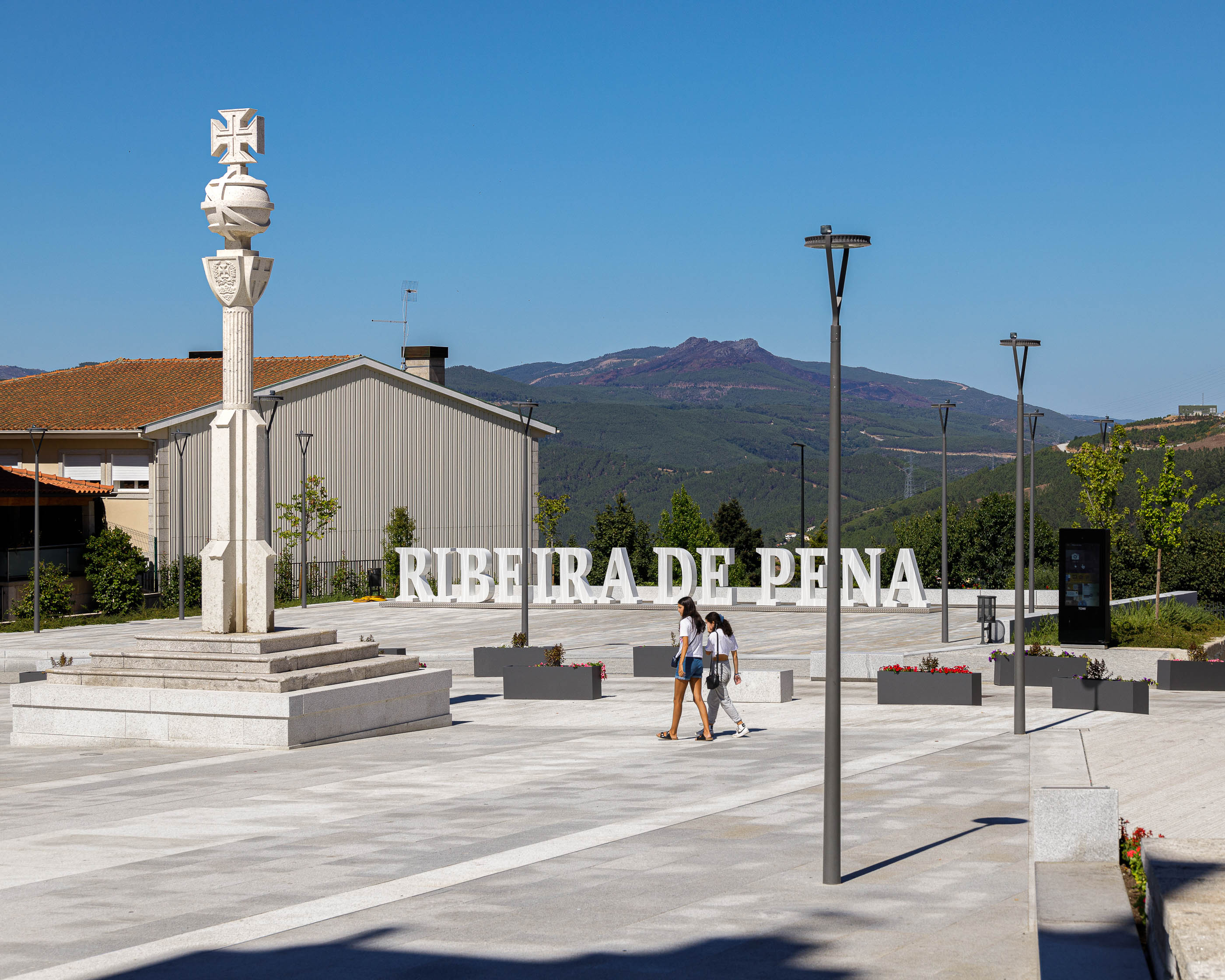 Praça: Cruzeiro em pedra, letras "RIBEIRA DE PENA", candeeiros modernos e pessoas a passear. — Requalificação da Praça do Município de Ribeira de Pena arquitetura AXR - Arquitetura e Design em Vila Real
