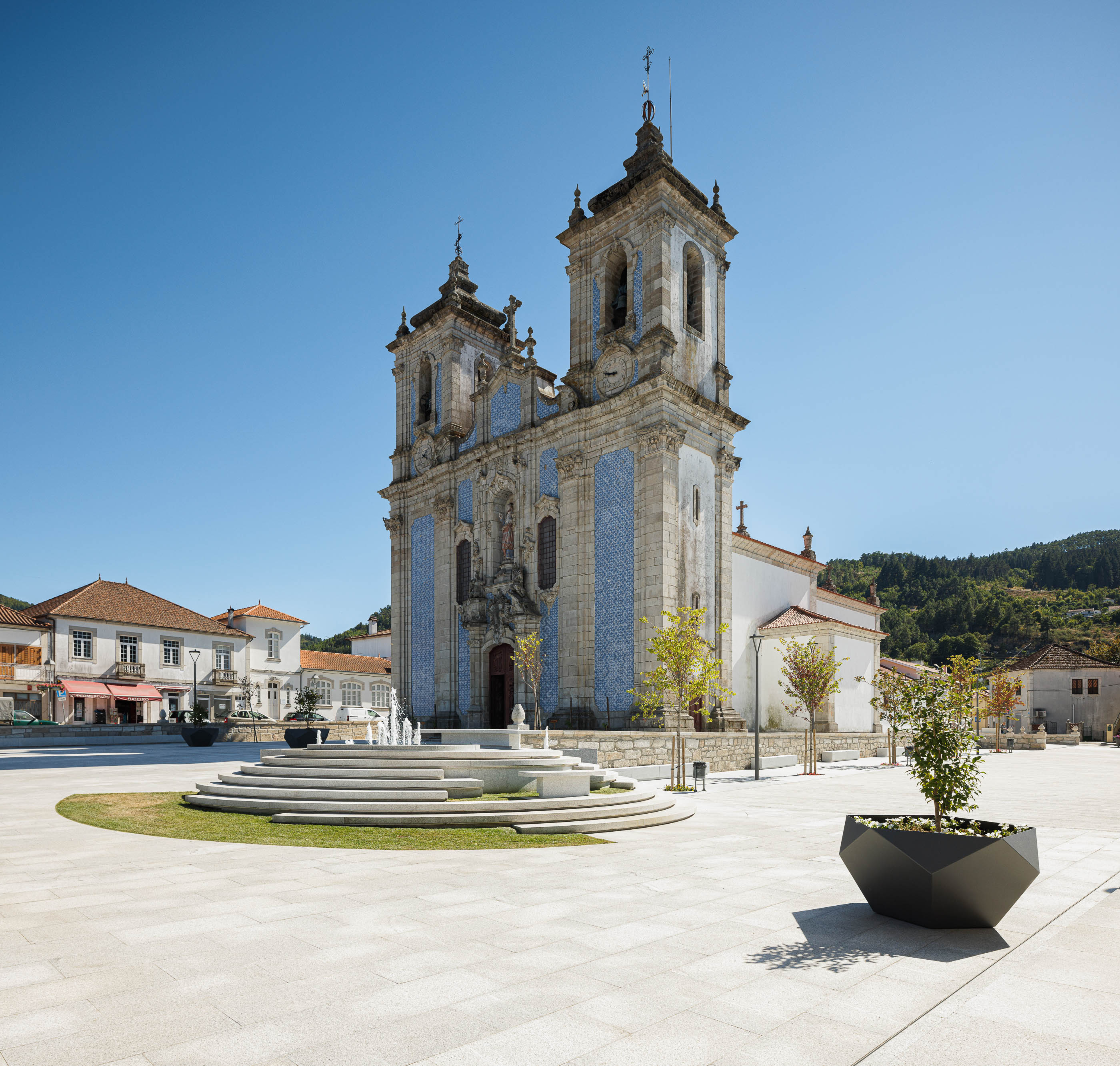 Praça: Igreja com fachada em azulejo azul e branco, fonte escalonada e pavimento em granito. — Requalificação da Praça do Município de Ribeira de Pena arquitetura AXR - Arquitetura e Design em Vila Real