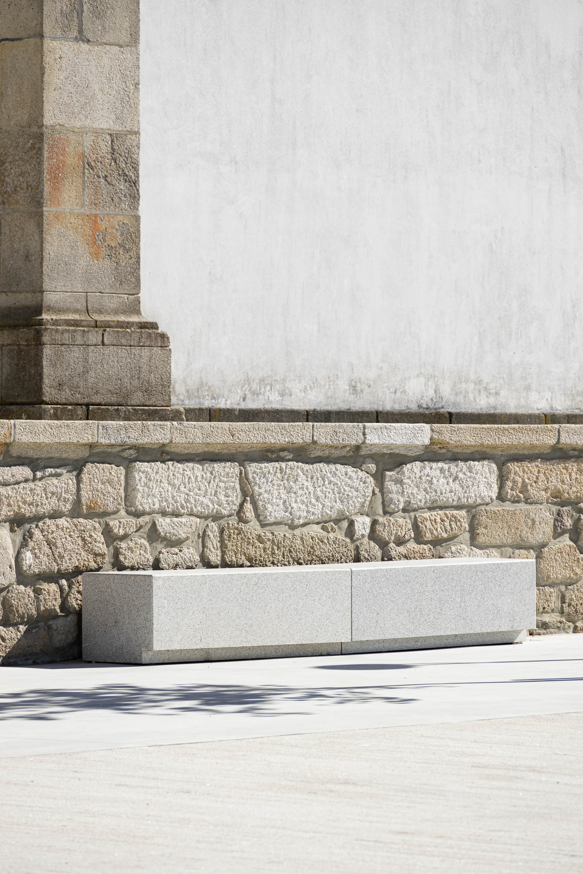 Praça: Banco em granito cinza claro encostado a muro de pedra rústica. — Requalificação da Praça do Município de Ribeira de Pena arquitetura AXR - Arquitetura e Design em Vila Real