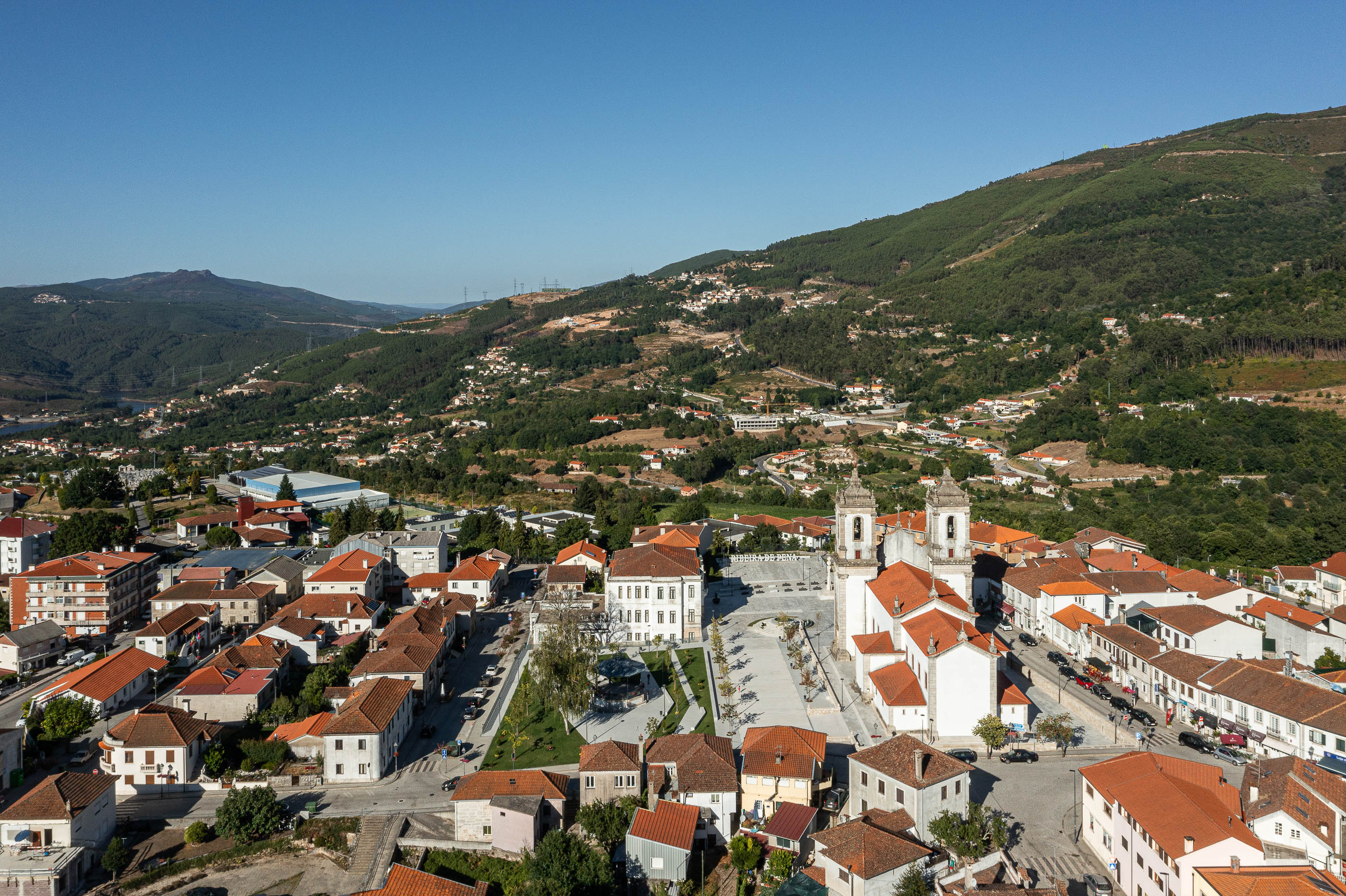 Praça: Vista aérea da vila com a praça central, igreja e edifícios com telhados de cor laranja. — Requalificação da Praça do Município de Ribeira de Pena arquitetura AXR - Arquitetura e Design em Vila Real