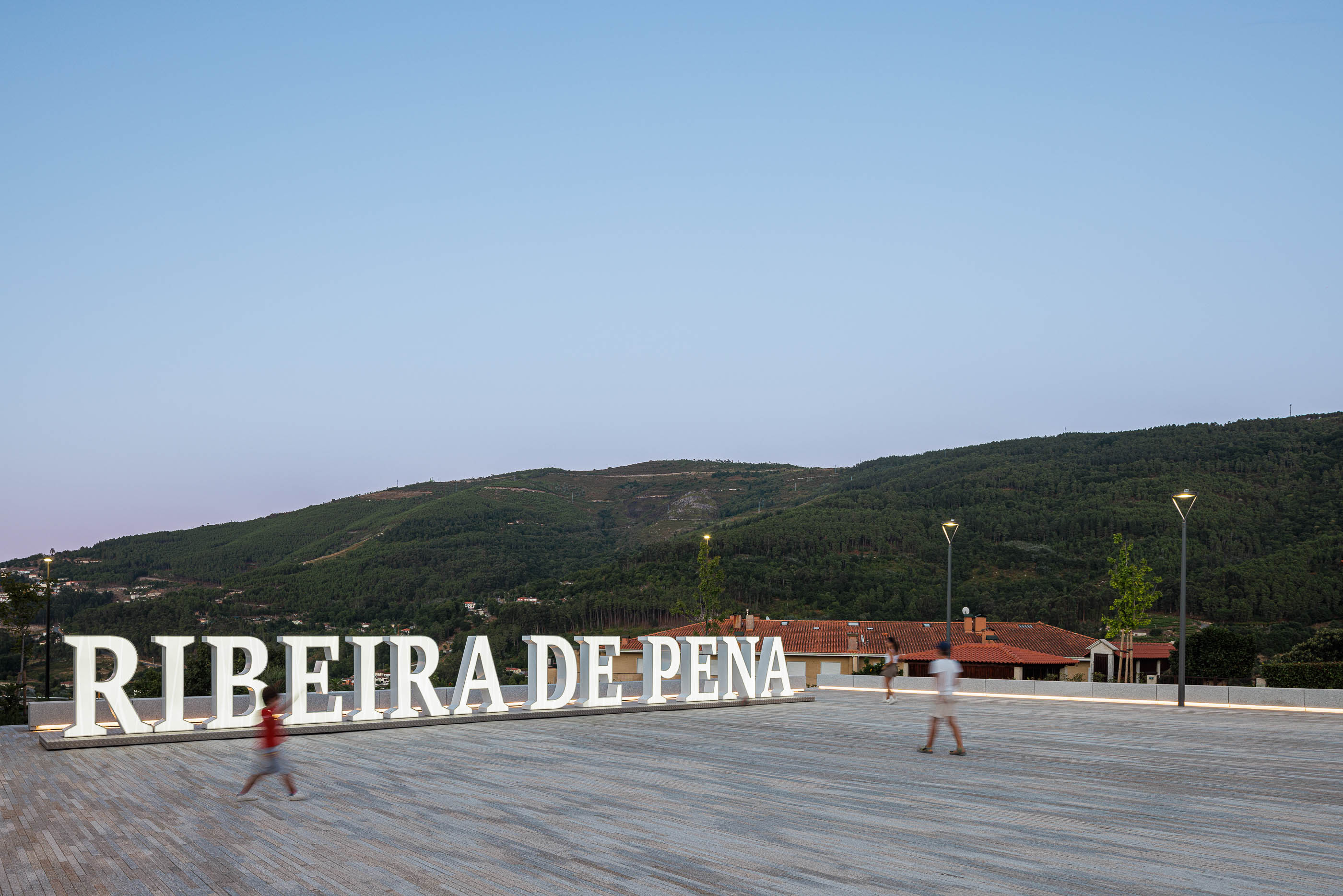 Praça: Letras "RIBEIRA DE PENA" iluminadas com pessoas a passear e montanhas ao fundo. — Requalificação da Praça do Município de Ribeira de Pena arquitetura AXR - Arquitetura e Design em Vila Real