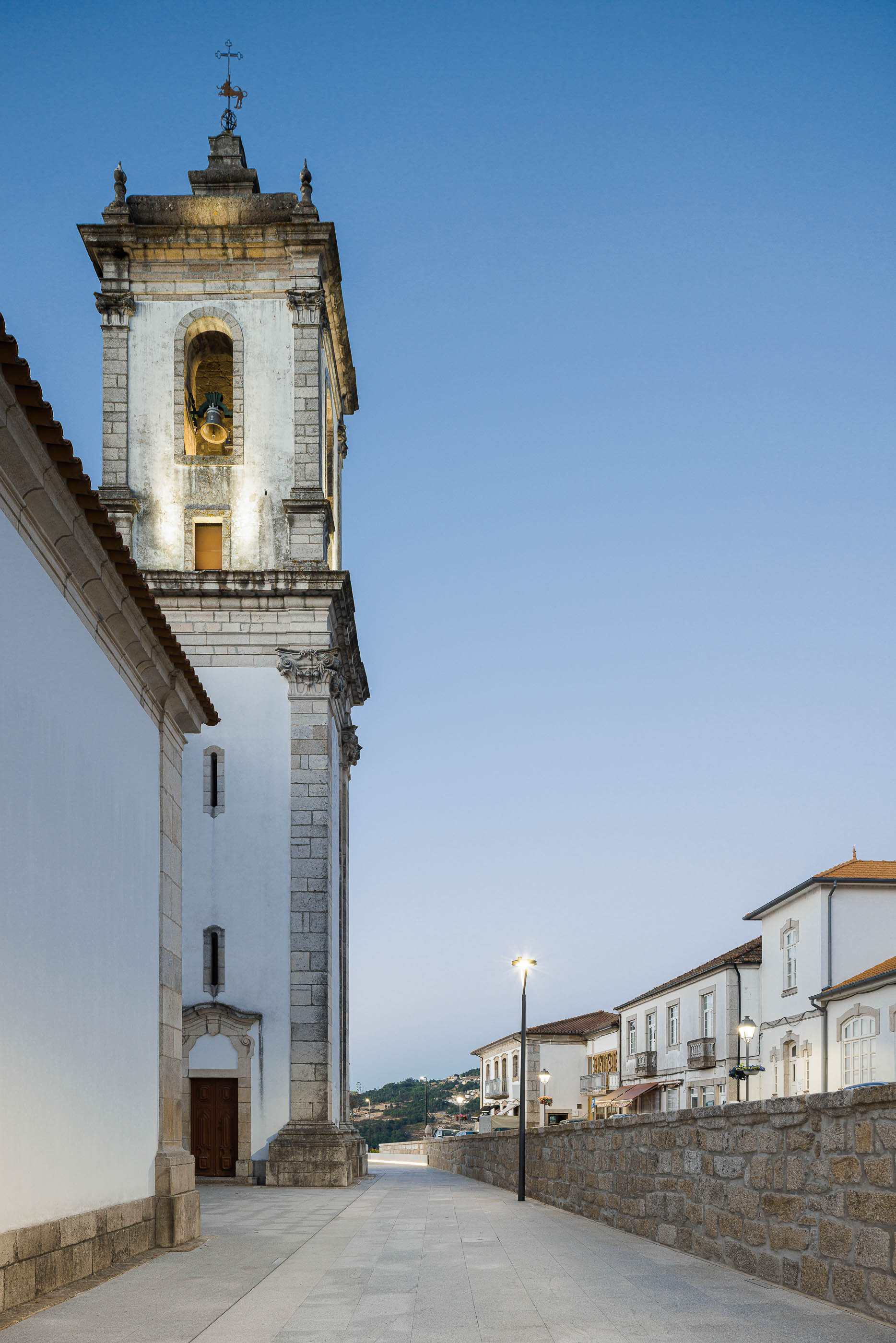 Rua: Torre sineira da igreja iluminada e rua com pavimento em granito. — Requalificação da Praça do Município de Ribeira de Pena arquitetura AXR - Arquitetura e Design em Vila Real