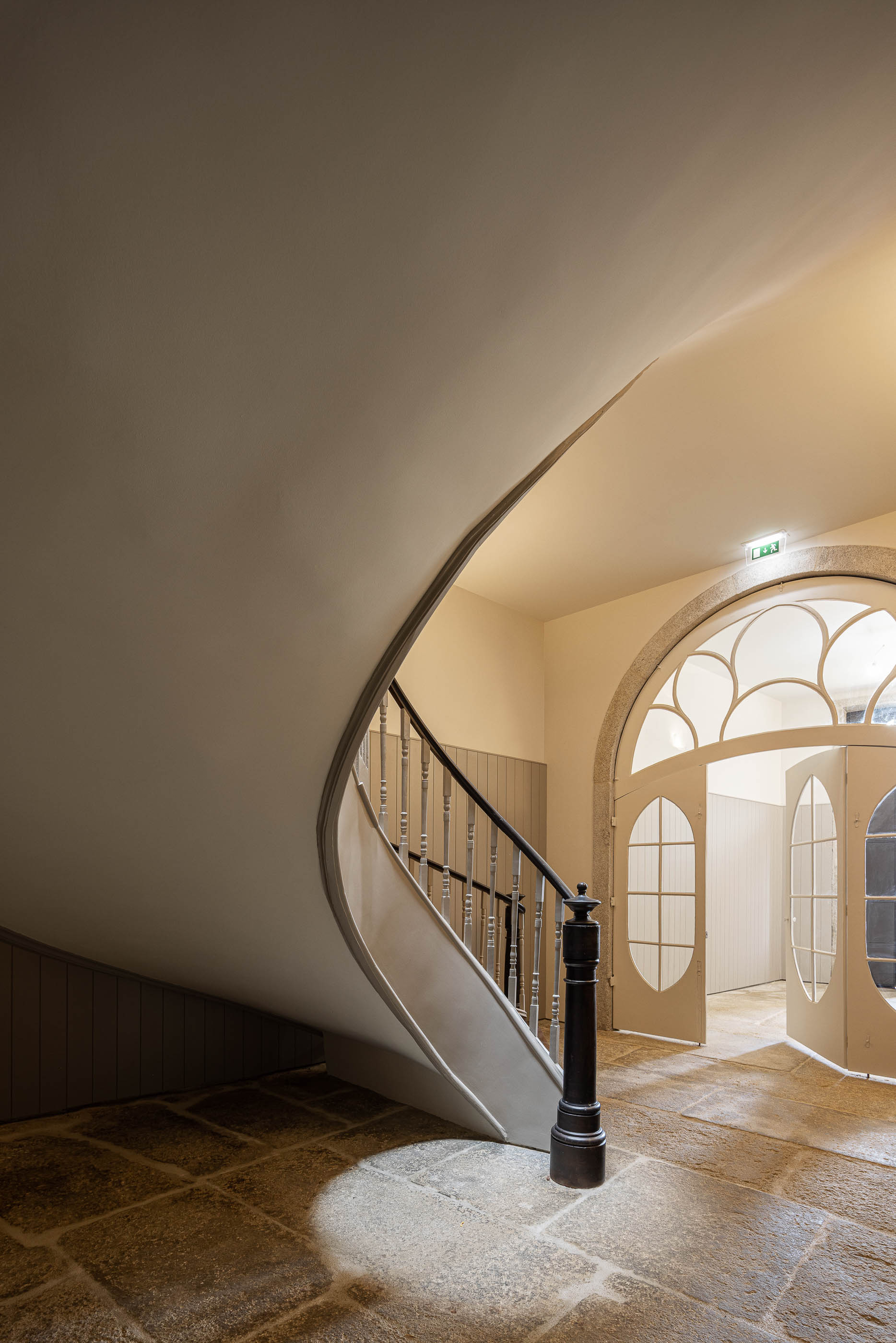 Escadaria: Detalhe da escadaria em caracol, com pavimento em pedra e porta em arco ao fundo. — Antigo Hotel do Louvre arquitetura Diana Barros Arquitectura em Porto