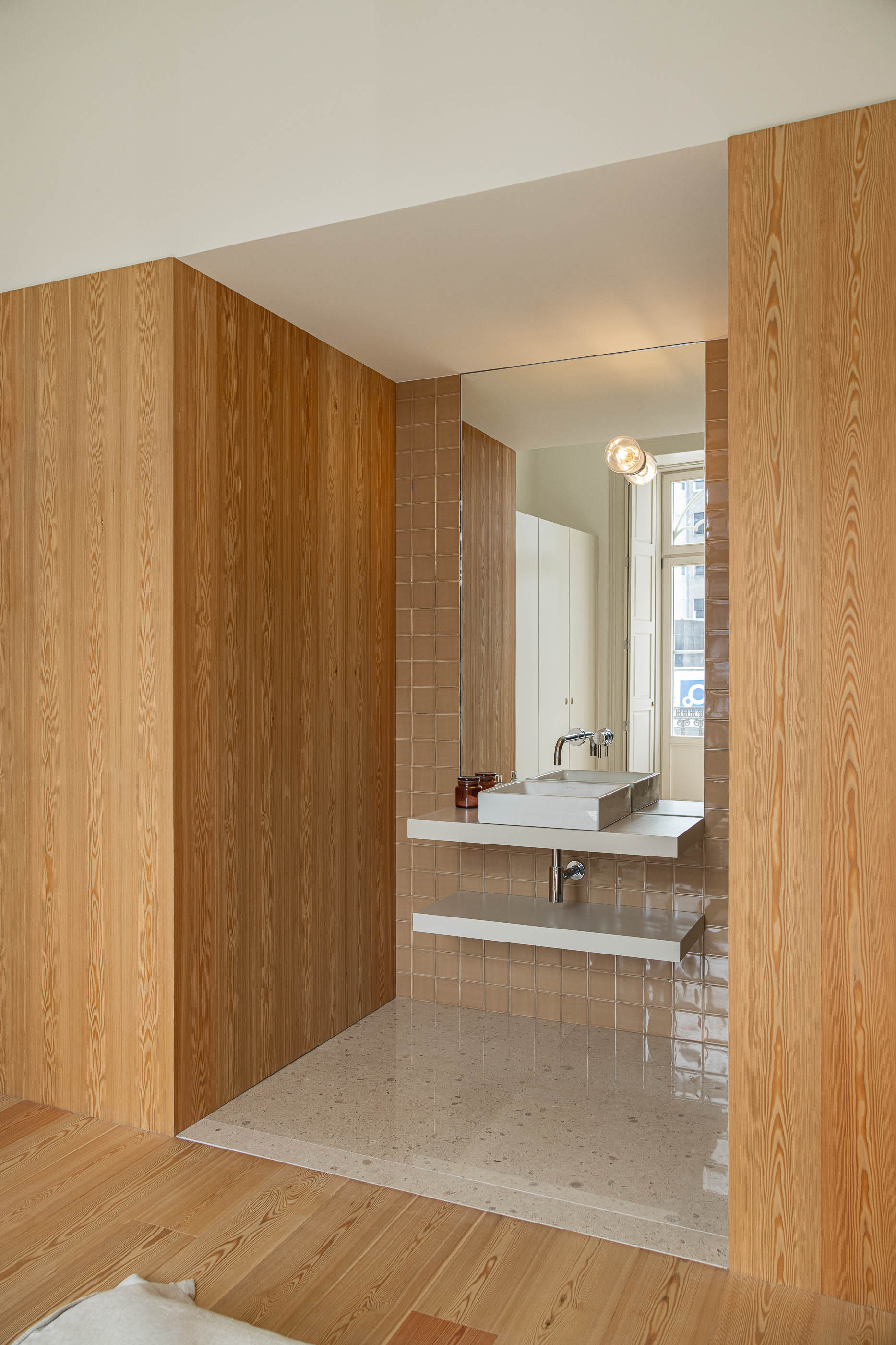Casa de banho: Casa de banho com revestimento em madeira clara, lavatório branco e espelho amplo. — Antigo Hotel do Louvre arquitetura Diana Barros Arquitectura em Porto
