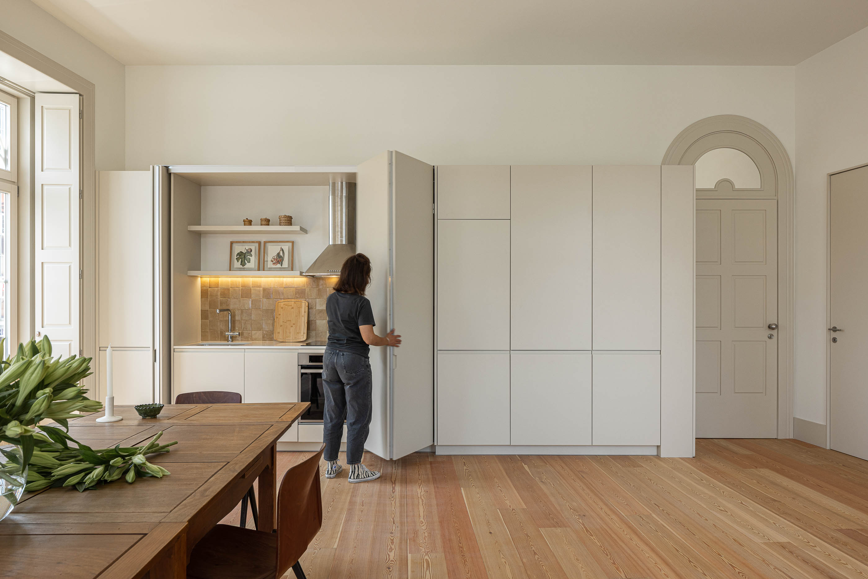 Sala de jantar: Mulher a abrir armário da cozinha moderna, com mesa de jantar em primeiro plano. — Antigo Hotel do Louvre arquitetura Diana Barros Arquitectura em Porto