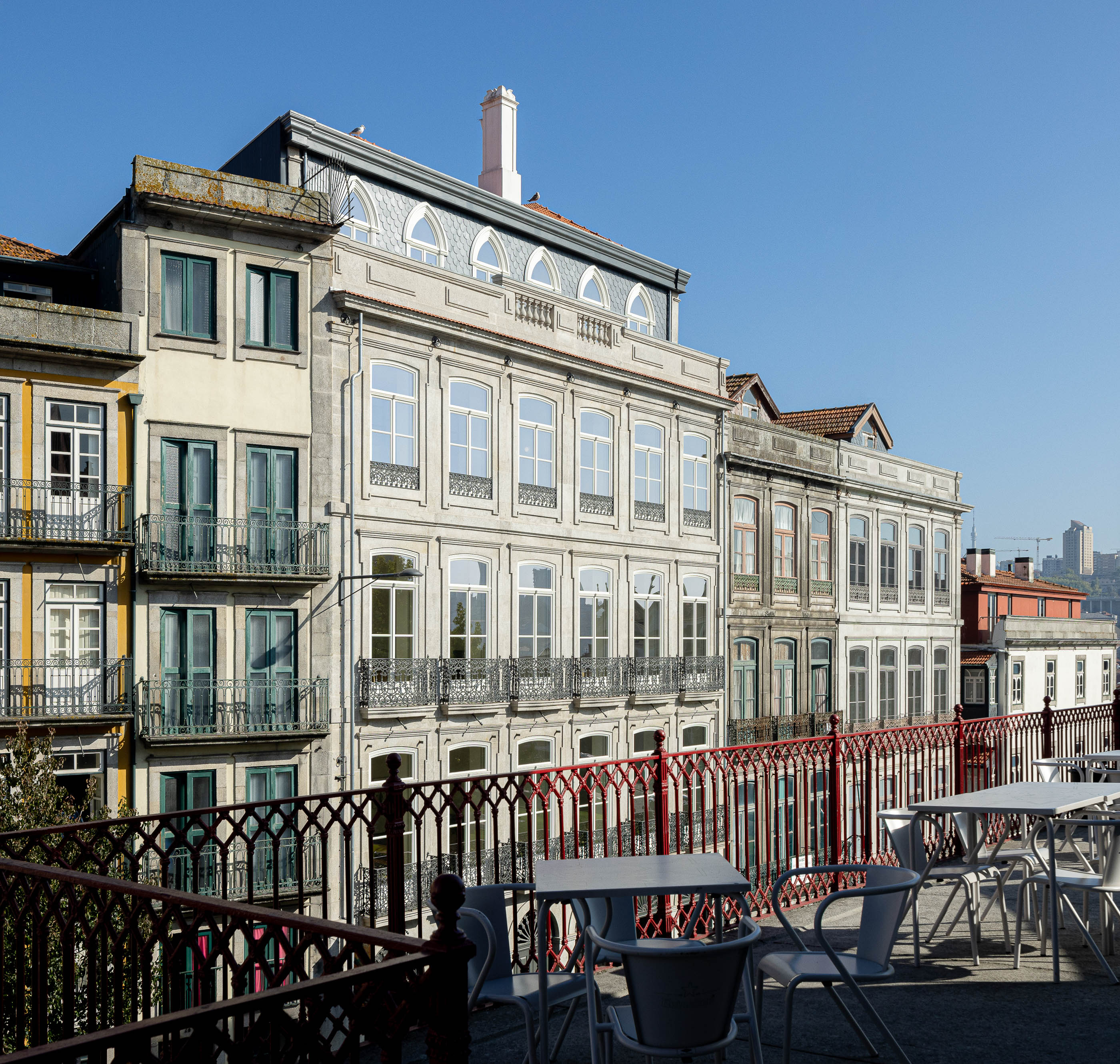 Terraço: Vista do edifício com mesas e cadeiras, gradeamento vermelho e edifícios históricos ao fundo. — Edifício de Mouzinho da Silveira arquitetura Diana Barros Arquitectura em Porto