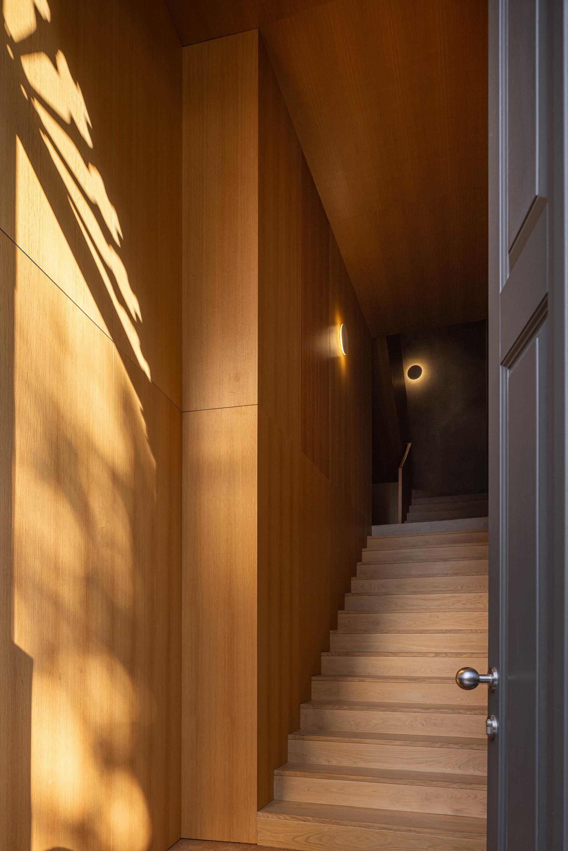 Entrada: Corredor com paredes em madeira clara, escadas em madeira e candeeiros de parede circulares. — Edifício de Mouzinho da Silveira arquitetura Diana Barros Arquitectura em Porto