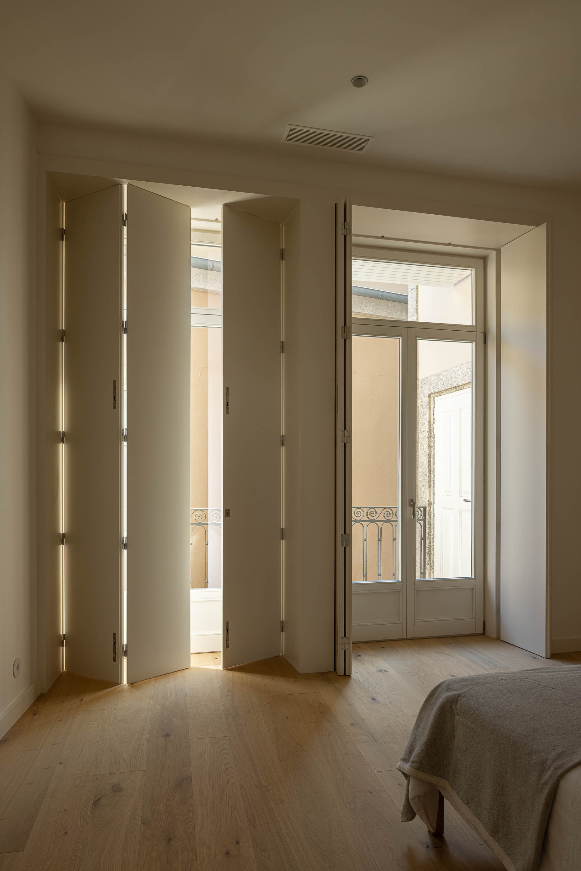 Quarto: Janela com portadas dobráveis brancas e pavimento em madeira clara. — Edifício de Mouzinho da Silveira arquitetura Diana Barros Arquitectura em Porto