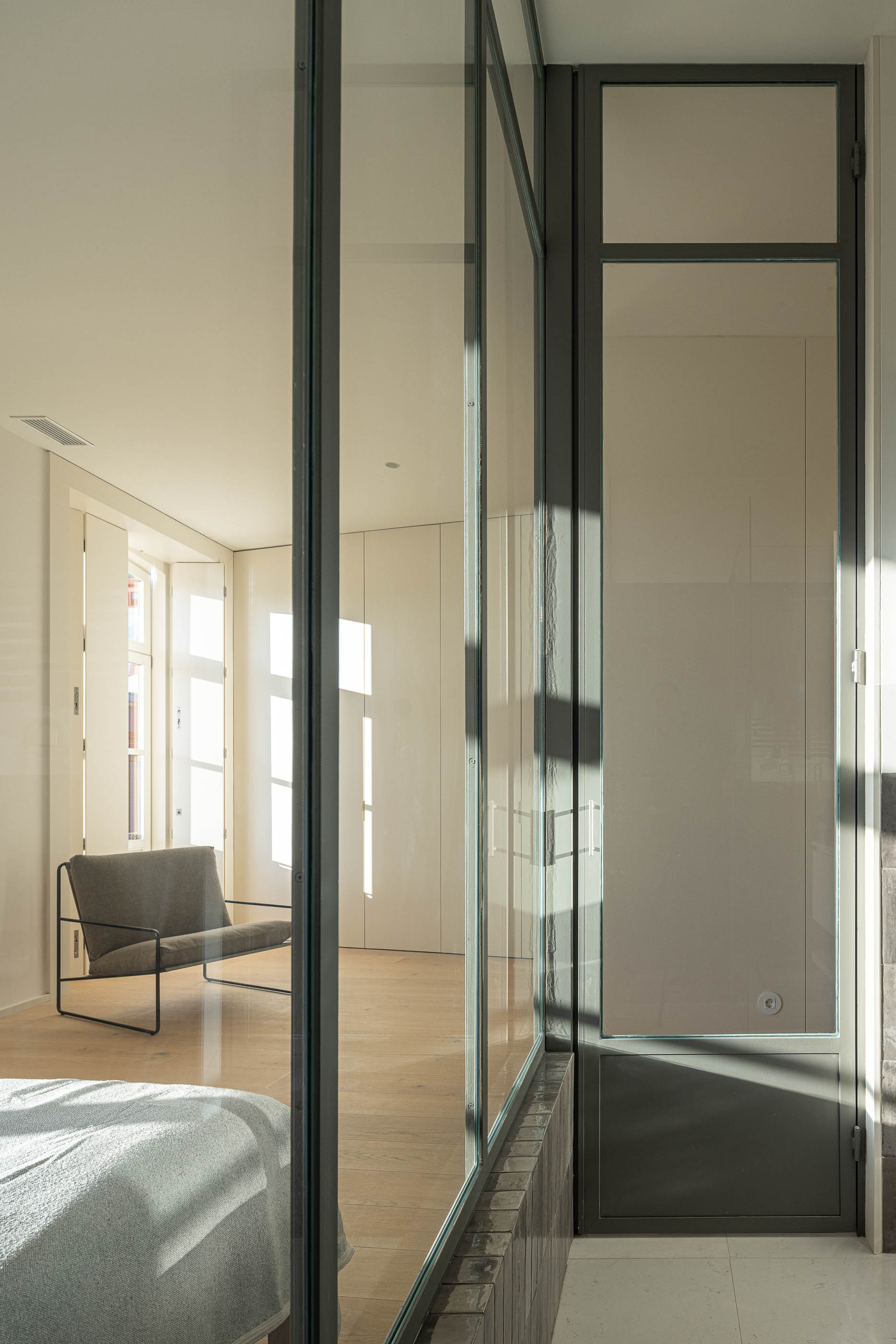 Quarto: Vista do quarto com casa de banho em vidro, roupeiros embutidos e cadeira. — Edifício de Mouzinho da Silveira arquitetura Diana Barros Arquitectura em Porto