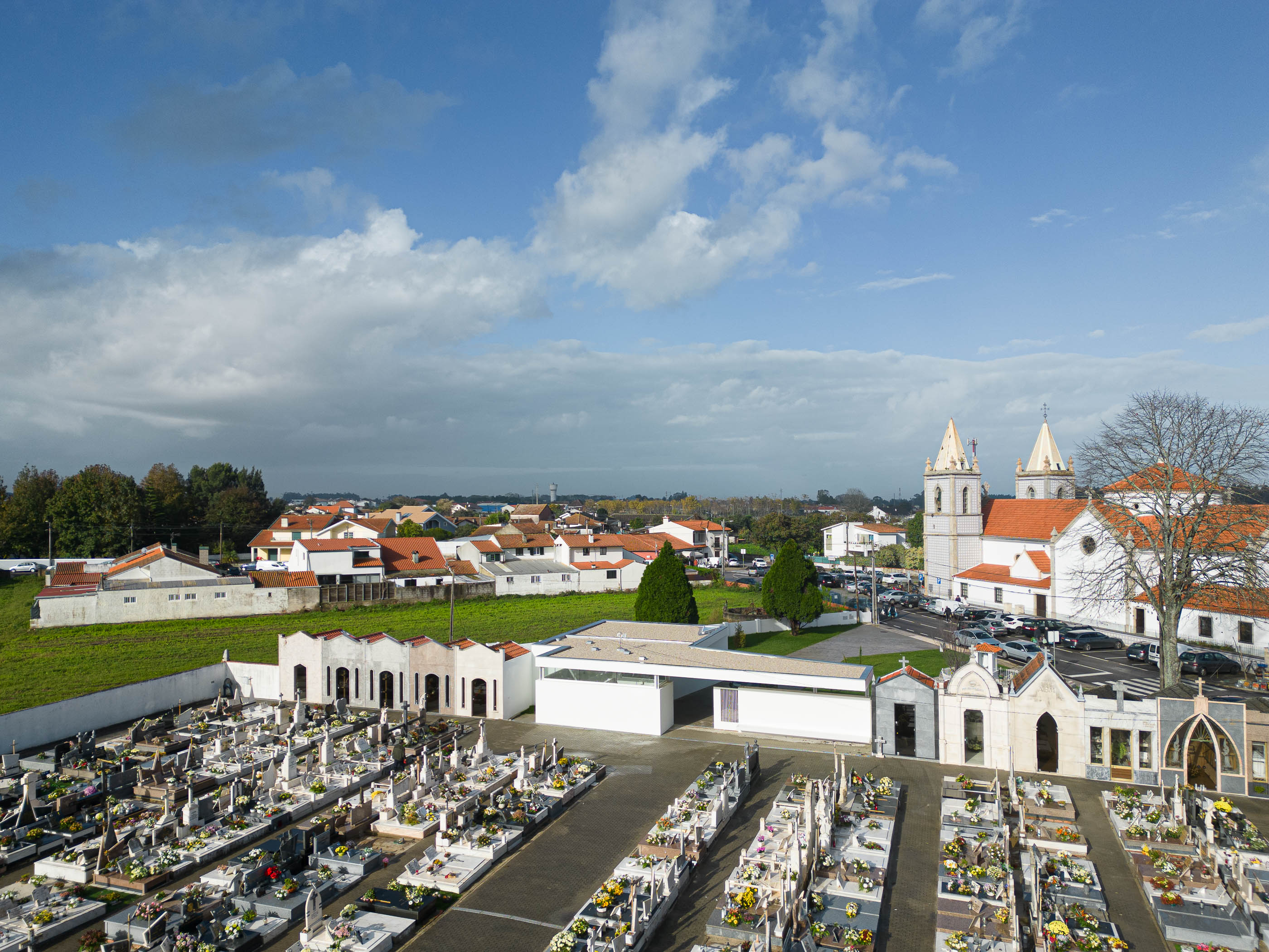 Vista geral: Cemitério com a casa mortuária branca de linhas modernas, a igreja e casas com telhados vermelhos ao fundo. — Casa Mortuária de Oliveirinha arquitetura Sónia Cruz Arquitectura em Aveiro