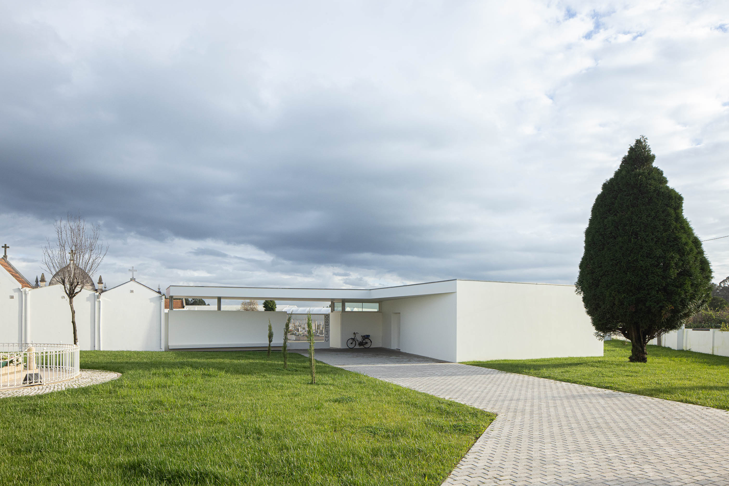 Exterior: Fachada branca da casa mortuária com alpendre, relvado e caminho em calçada portuguesa. — Casa Mortuária de Oliveirinha arquitetura Sónia Cruz Arquitectura em Aveiro