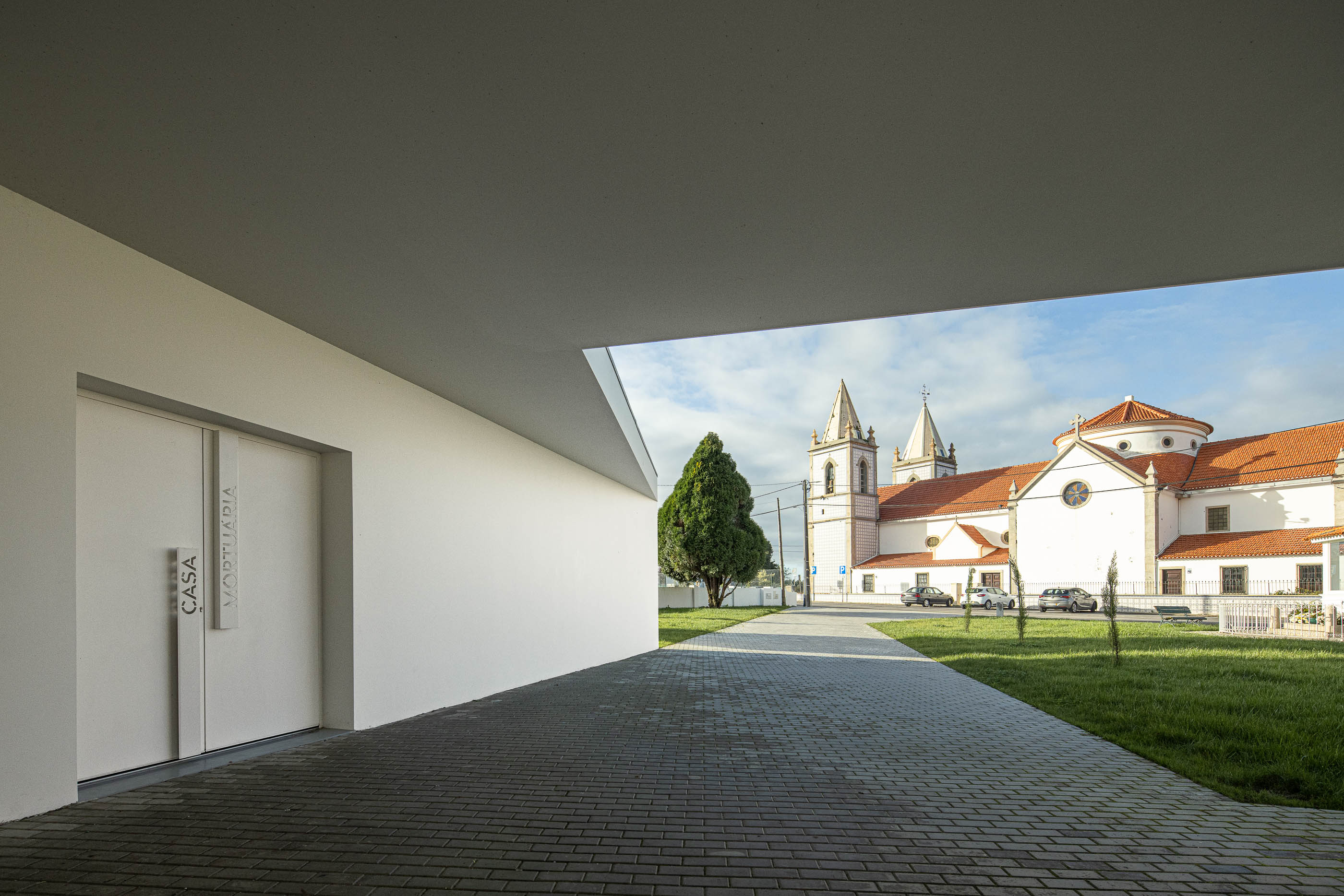 Exterior: Entrada da casa mortuária com porta dupla e vista para a igreja ao fundo. — Casa Mortuária de Oliveirinha arquitetura Sónia Cruz Arquitectura em Aveiro