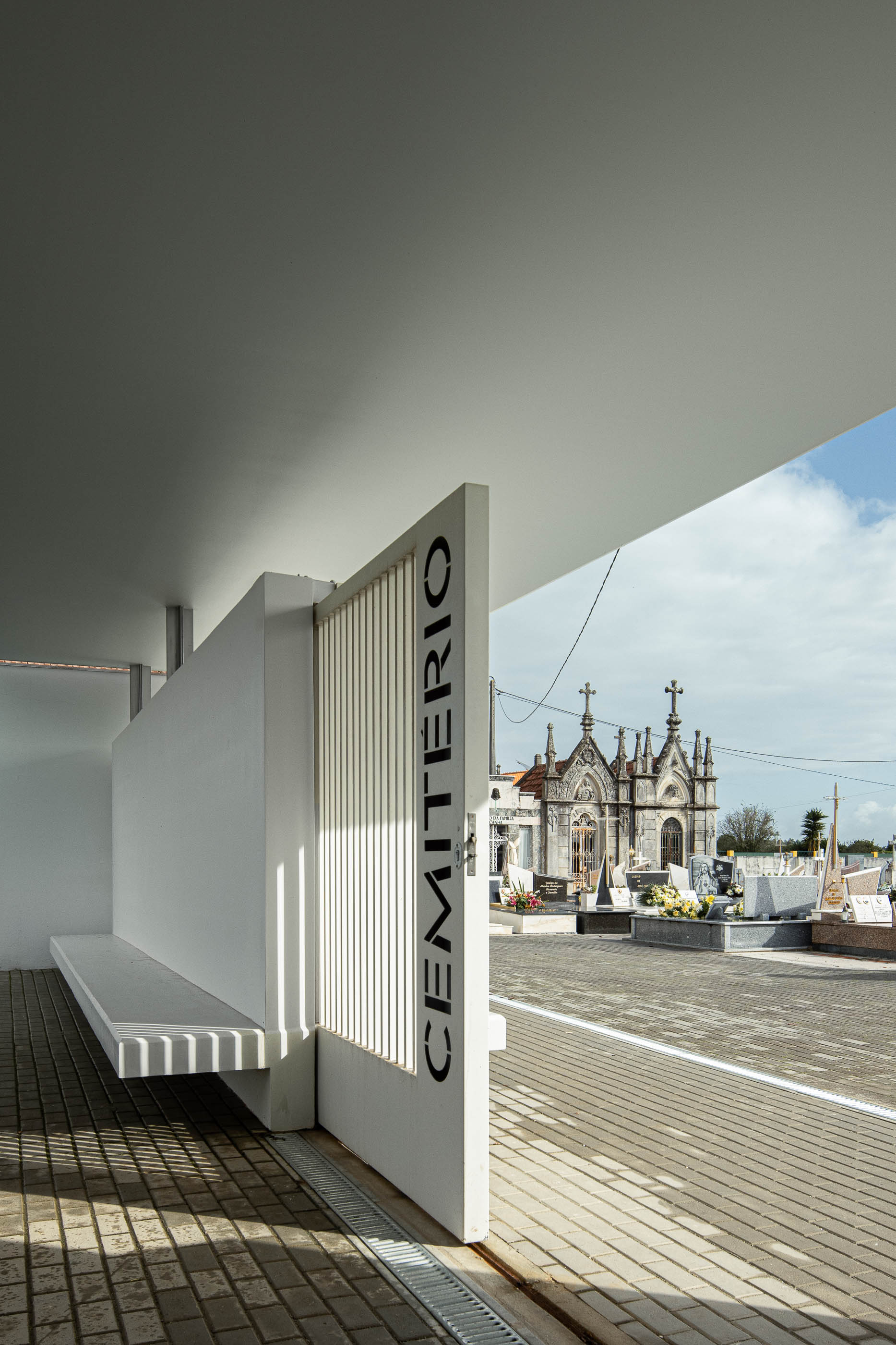 Exterior: Alpendre da casa mortuária com banco embutido e porta gradeada para o cemitério. — Casa Mortuária de Oliveirinha arquitetura Sónia Cruz Arquitectura em Aveiro