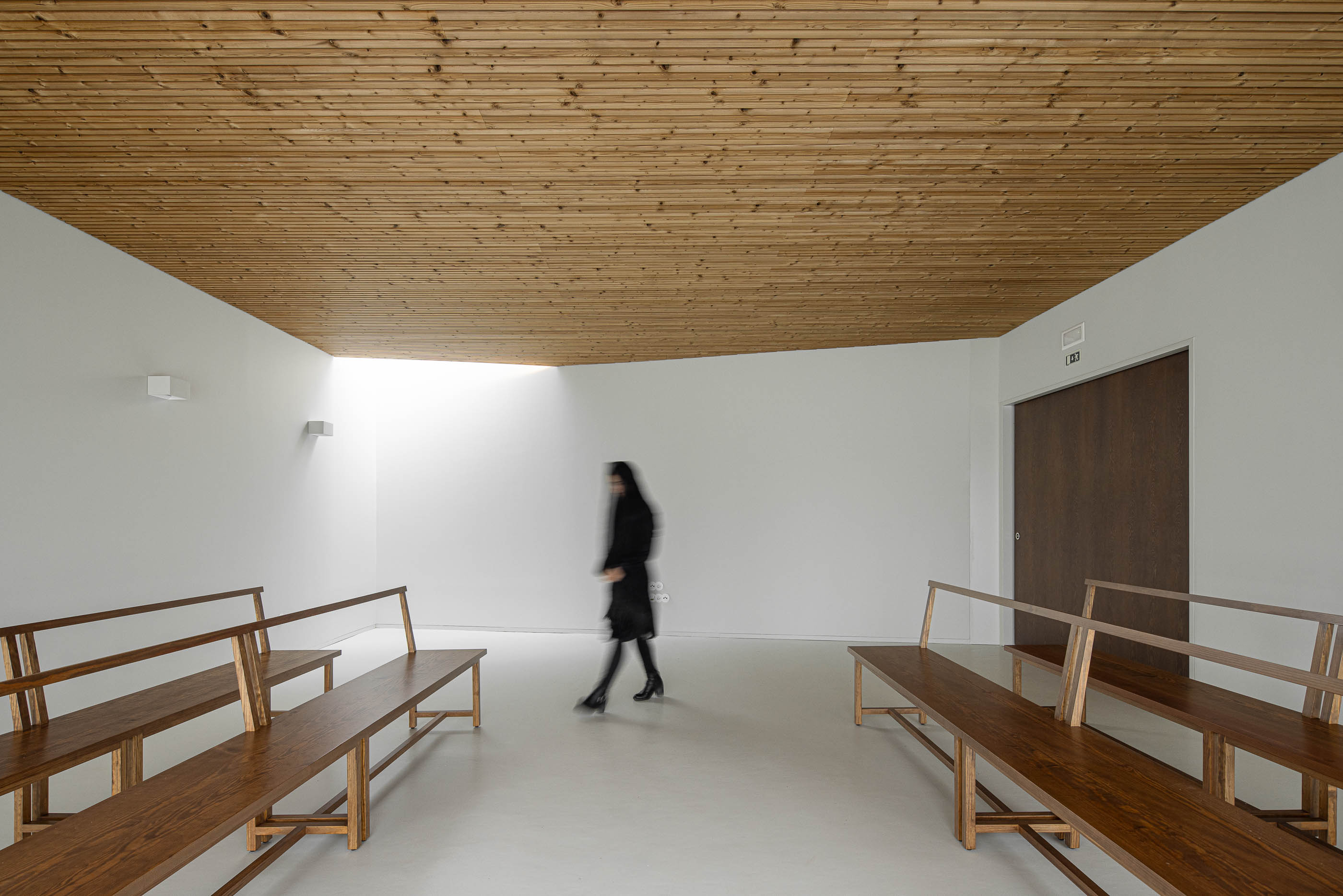Interior: Sala de cerimónias com teto em madeira, pavimento claro e bancos de madeira. — Casa Mortuária de Oliveirinha arquitetura Sónia Cruz Arquitectura em Aveiro