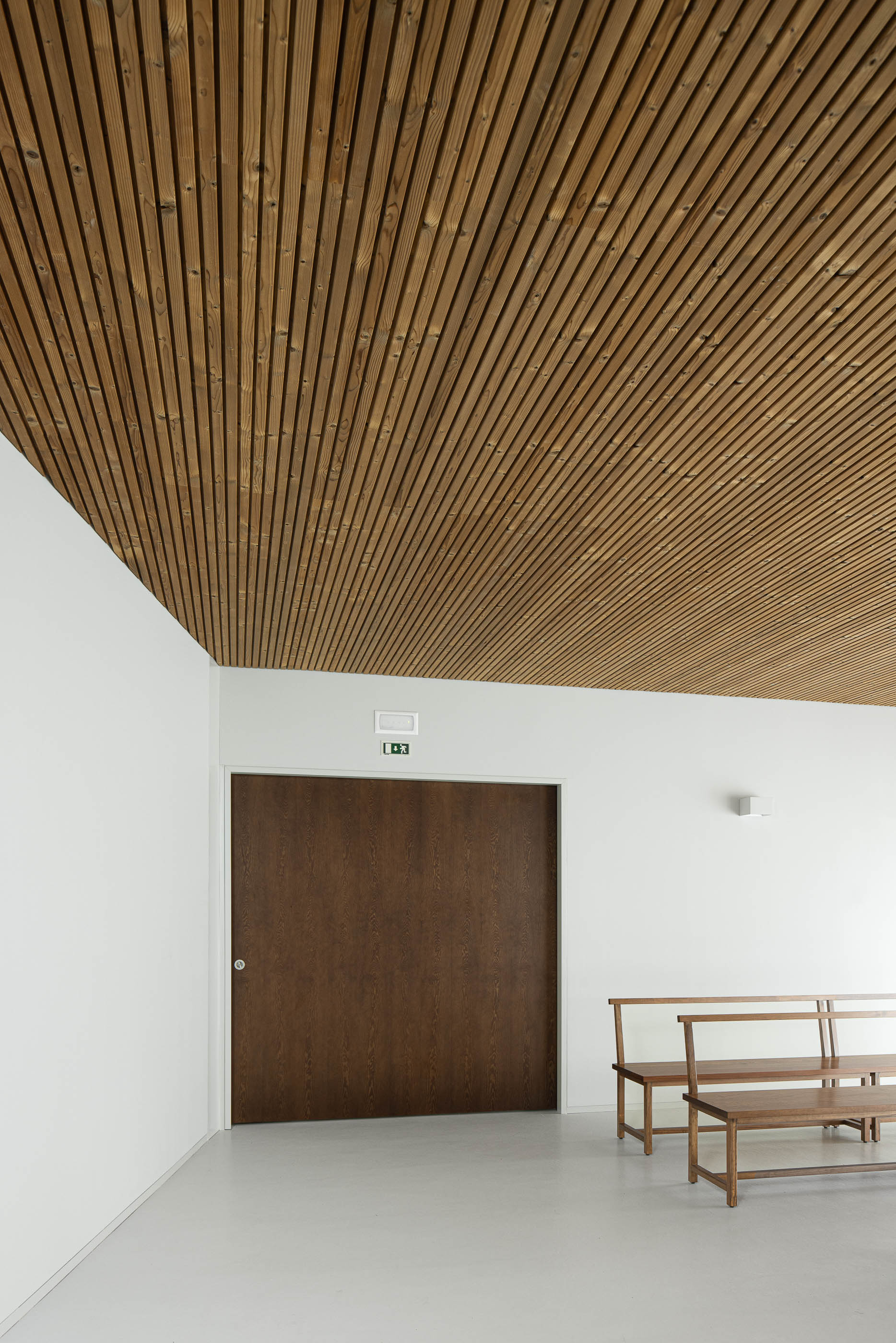 Interior: Sala de cerimónias com teto em madeira, porta de madeira e bancos de madeira. — Casa Mortuária de Oliveirinha arquitetura Sónia Cruz Arquitectura em Aveiro