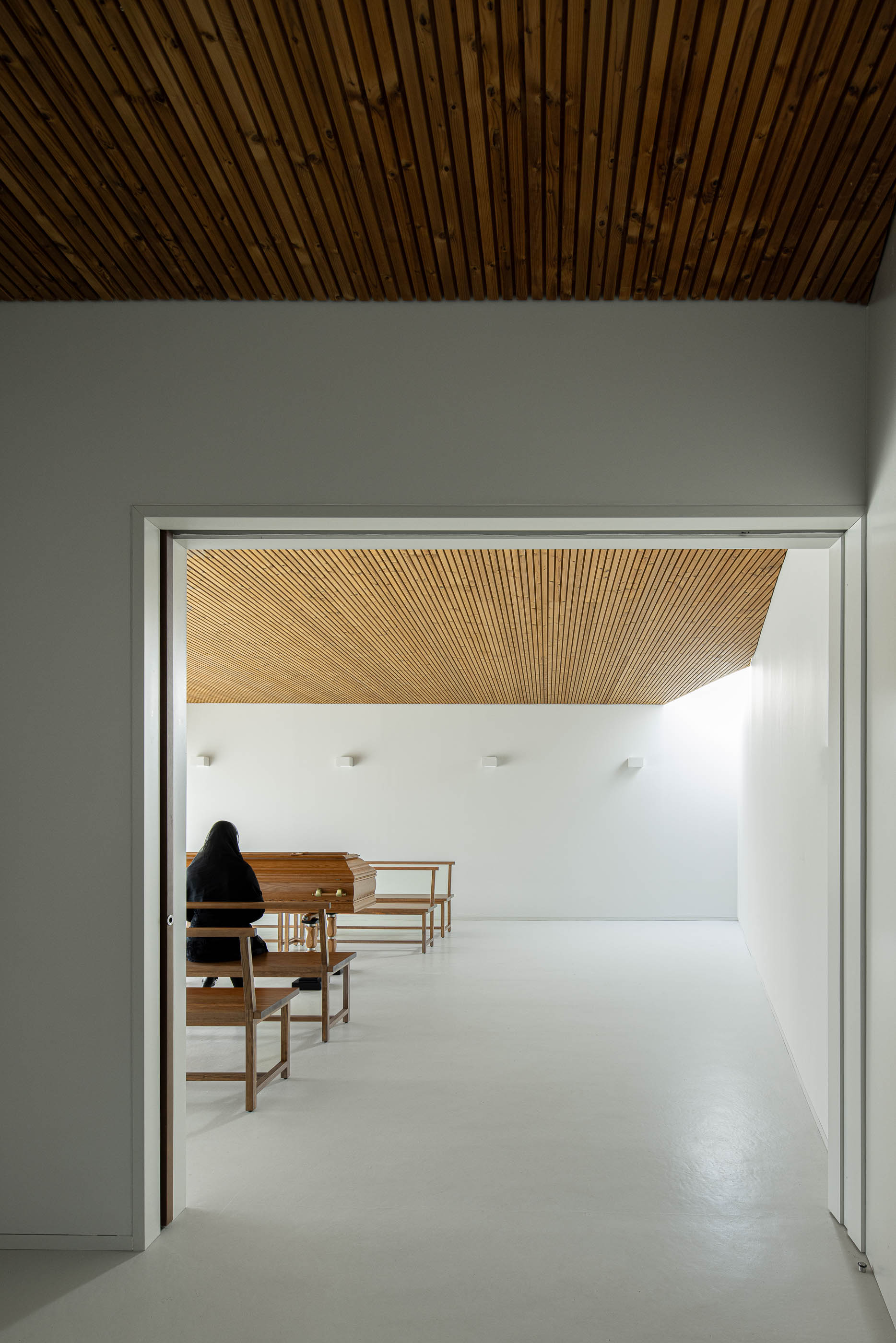Interior: Vista da sala de cerimónias com caixão, através de uma porta. Teto em madeira. — Casa Mortuária de Oliveirinha arquitetura Sónia Cruz Arquitectura em Aveiro