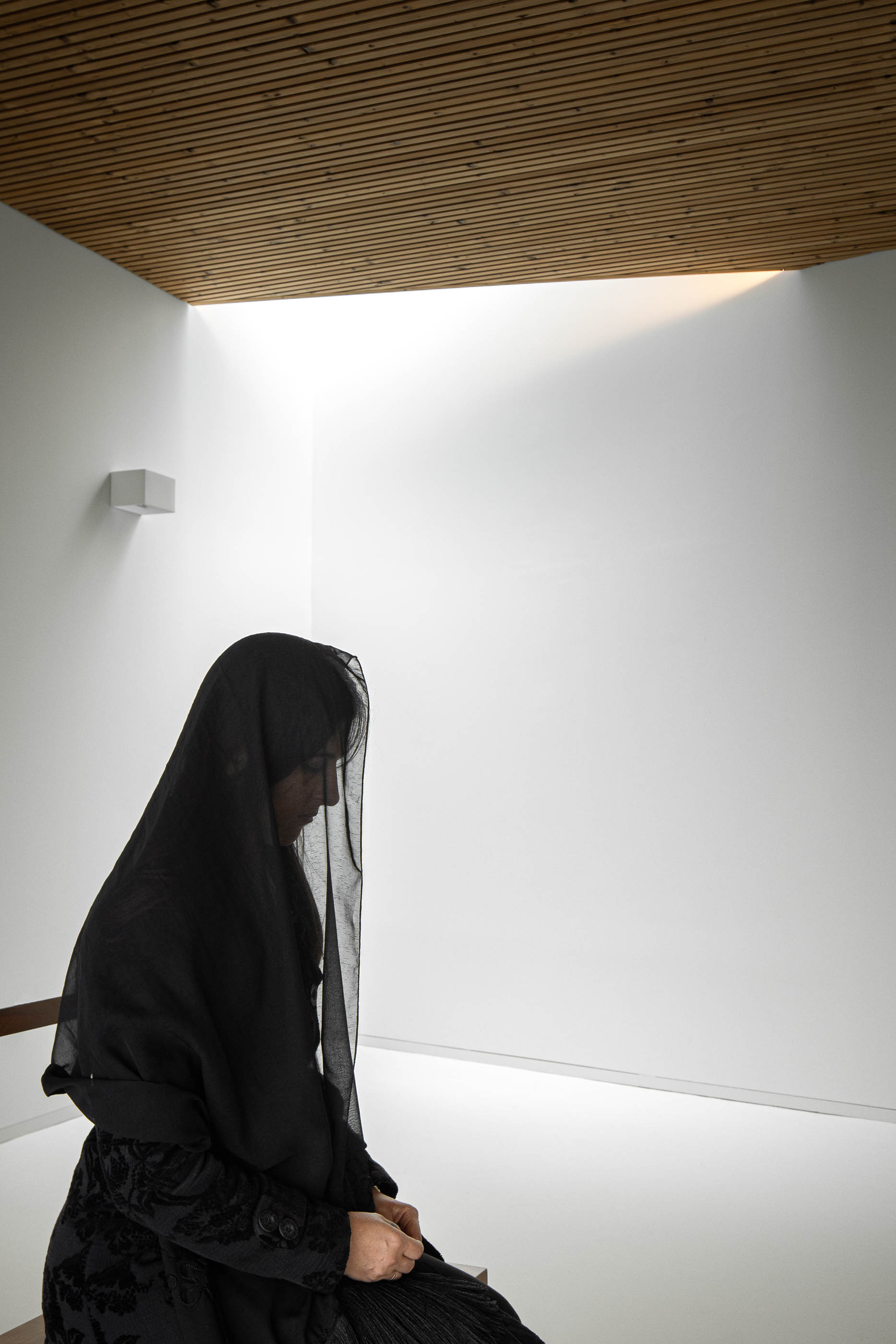 Interior: Pessoa sentada num banco na sala de cerimónias, com teto em madeira e luz zenital. — Casa Mortuária de Oliveirinha arquitetura Sónia Cruz Arquitectura em Aveiro