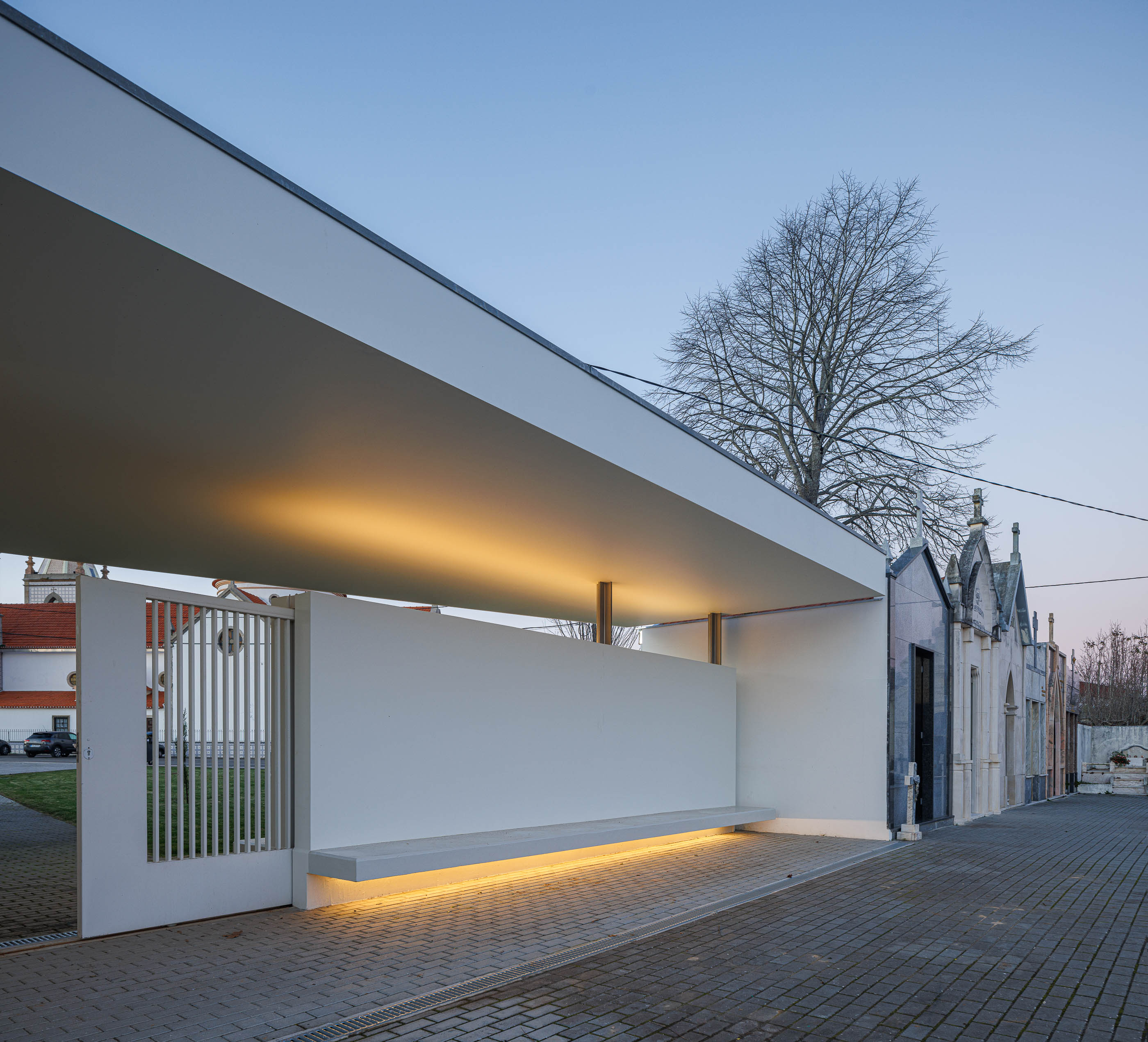 Exterior: Alpendre da casa mortuária com iluminação indireta e porta gradeada para o cemitério. — Casa Mortuária de Oliveirinha arquitetura Sónia Cruz Arquitectura em Aveiro