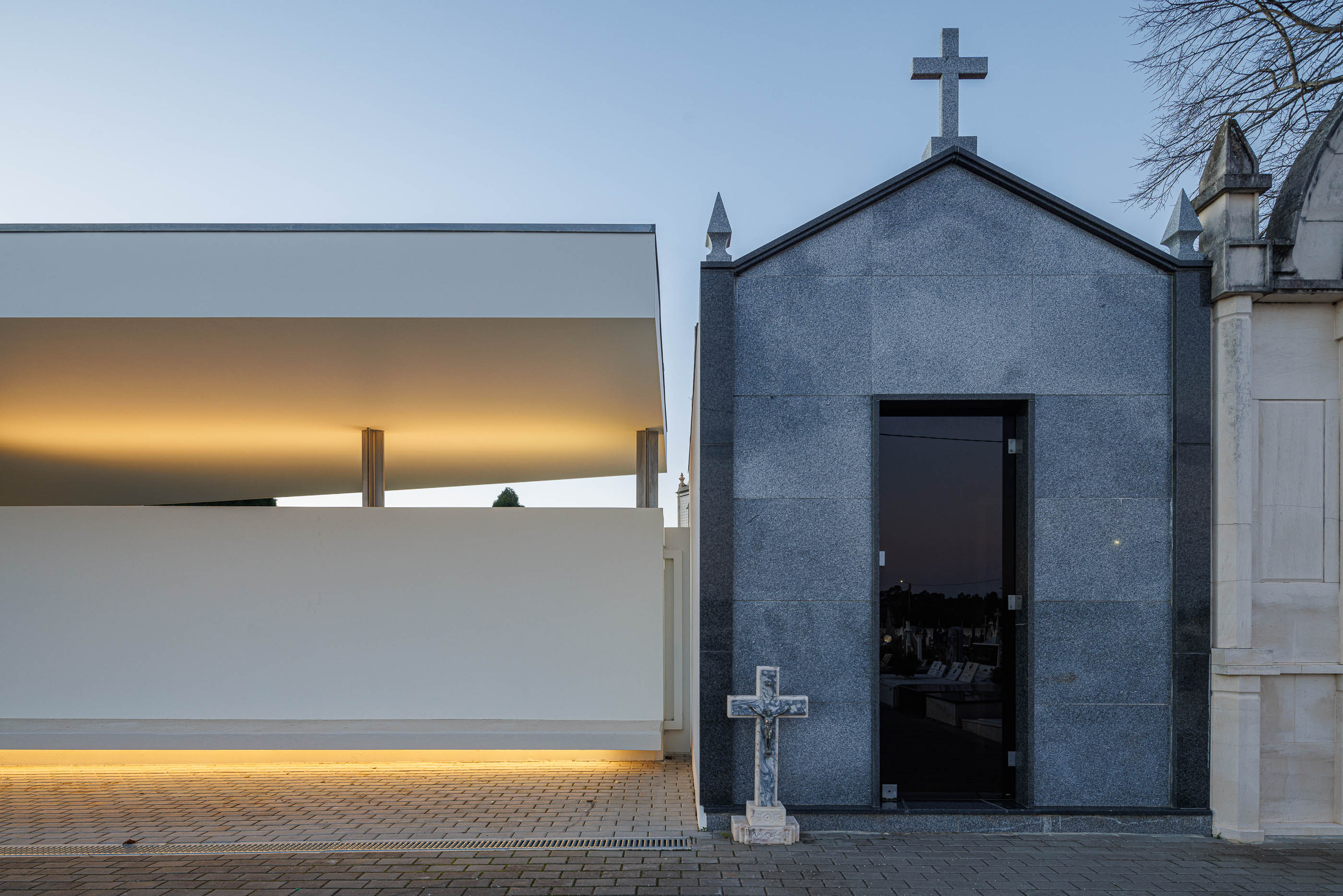 Exterior: Casa mortuária branca com alpendre iluminado, junto a uma capela em granito. — Casa Mortuária de Oliveirinha arquitetura Sónia Cruz Arquitectura em Aveiro