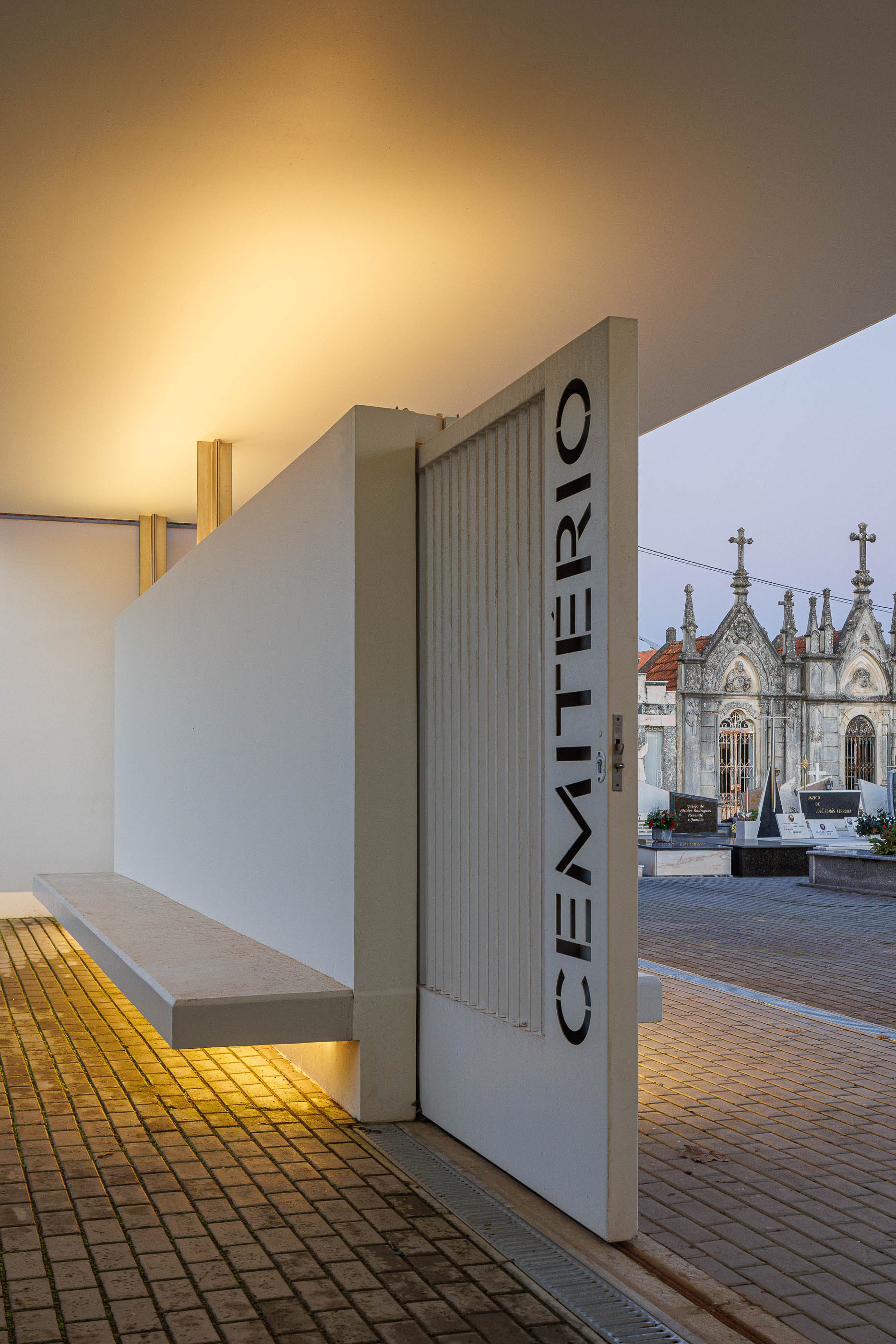 Exterior: Porta gradeada do alpendre da casa mortuária para o cemitério, com iluminação indireta. — Casa Mortuária de Oliveirinha arquitetura Sónia Cruz Arquitectura em Aveiro