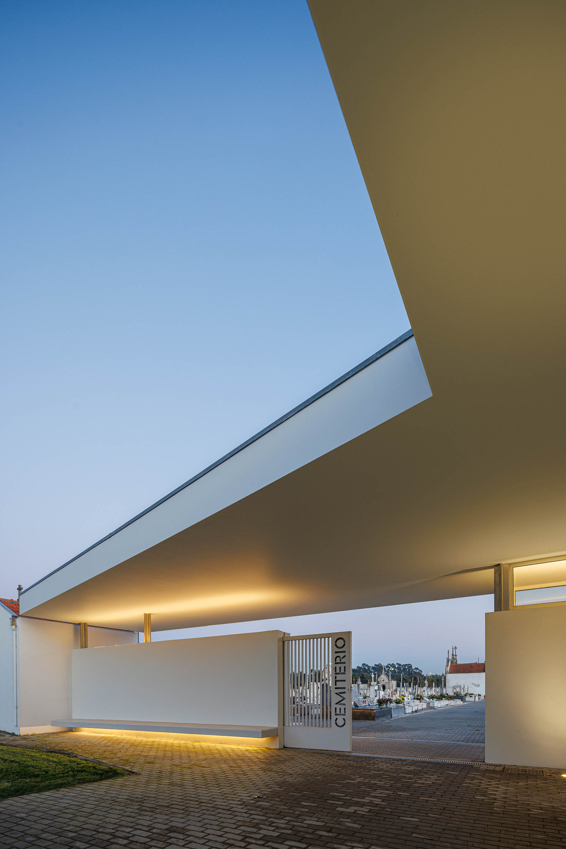 Exterior: Alpendre da casa mortuária iluminado, com porta gradeada para o cemitério. — Casa Mortuária de Oliveirinha arquitetura Sónia Cruz Arquitectura em Aveiro