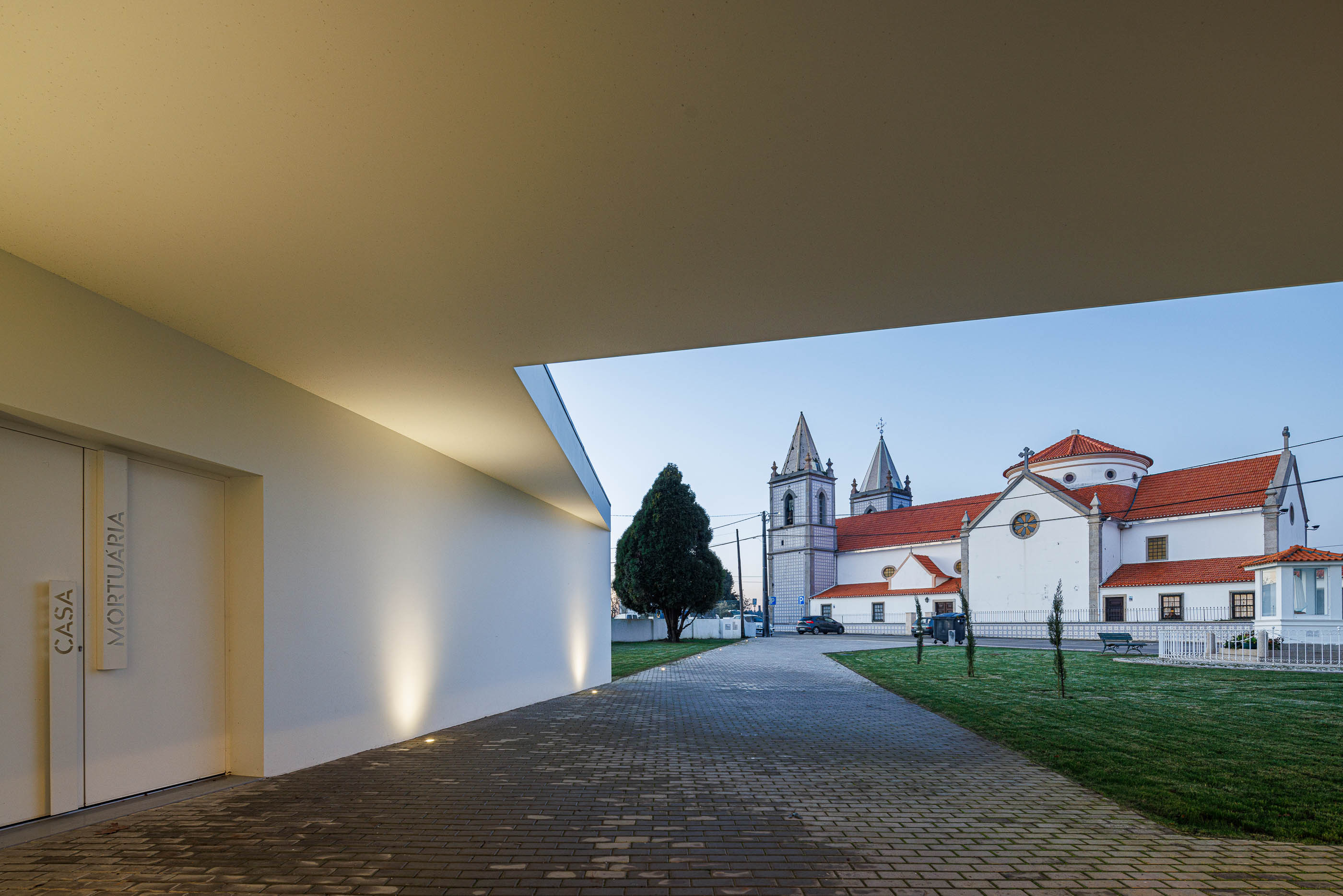 Exterior: Entrada da casa mortuária com porta dupla e vista para a igreja iluminada. — Casa Mortuária de Oliveirinha arquitetura Sónia Cruz Arquitectura em Aveiro