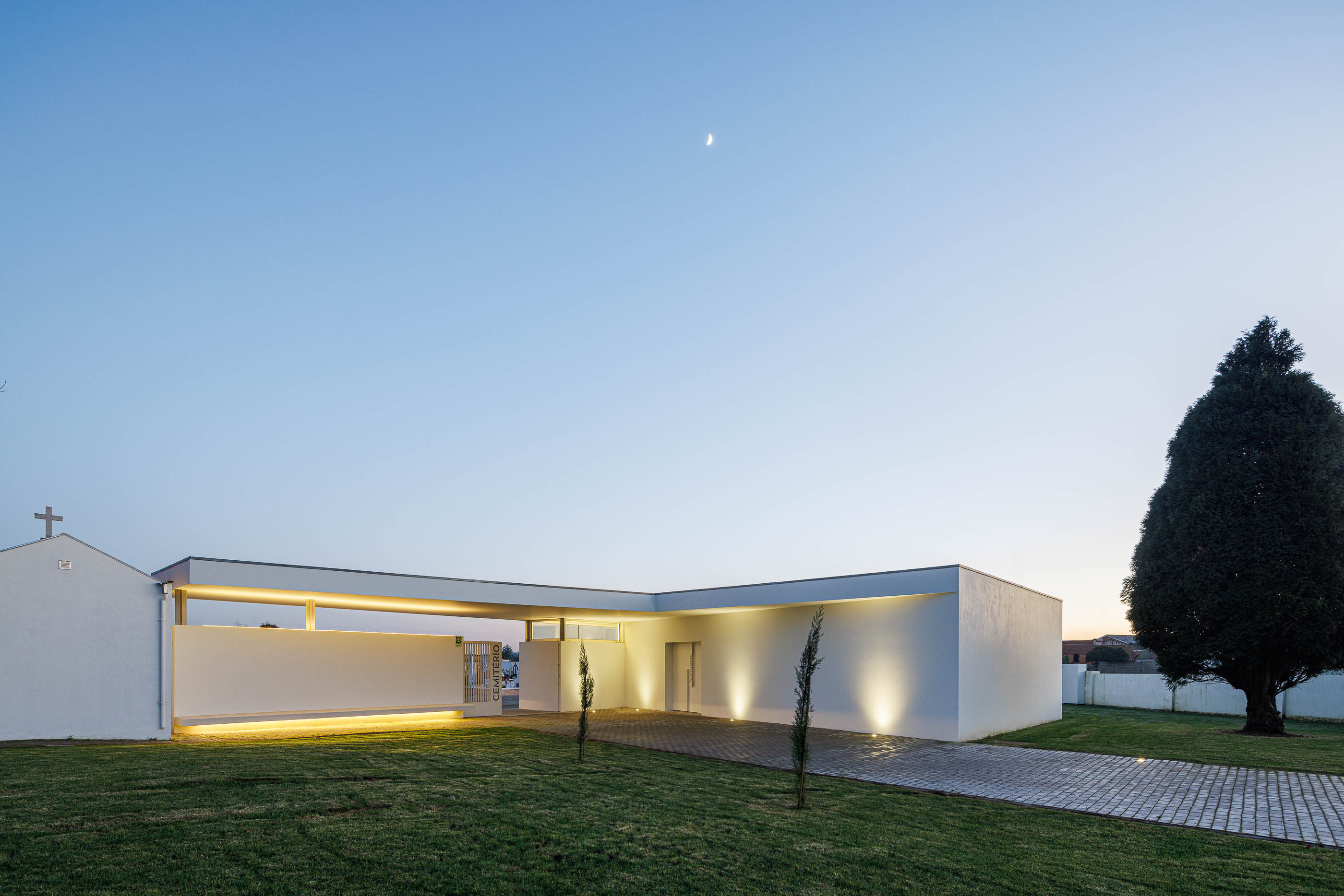 Exterior: Vista noturna da casa mortuária branca com alpendre iluminado e relvado. — Casa Mortuária de Oliveirinha arquitetura Sónia Cruz Arquitectura em Aveiro