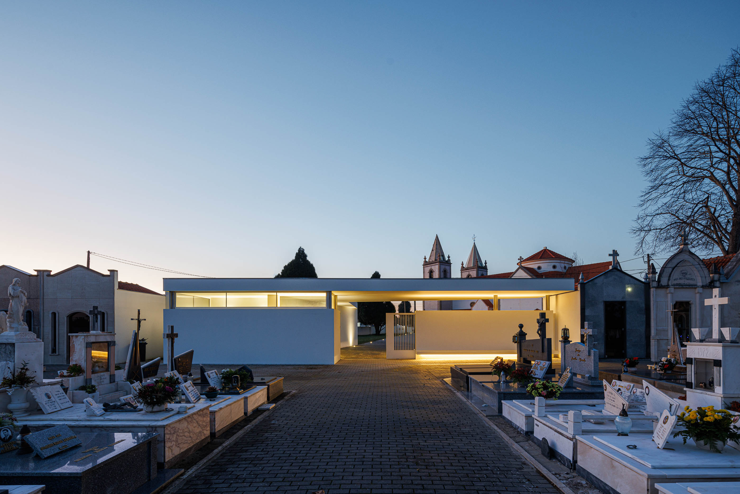 Exterior: Vista noturna da casa mortuária branca com alpendre iluminado, vista do cemitério. — Casa Mortuária de Oliveirinha arquitetura Sónia Cruz Arquitectura em Aveiro