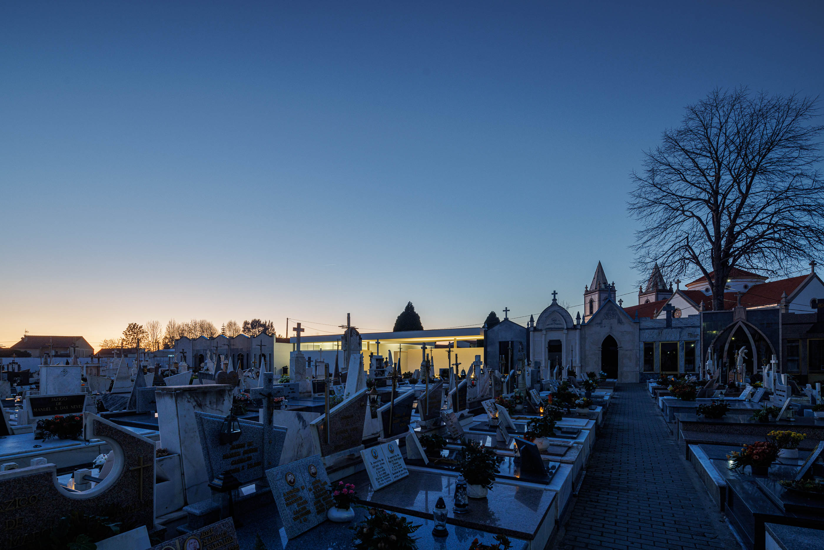 Vista geral: Cemitério com a casa mortuária branca iluminada e a igreja ao fundo, ao anoitecer. — Casa Mortuária de Oliveirinha arquitetura Sónia Cruz Arquitectura em Aveiro