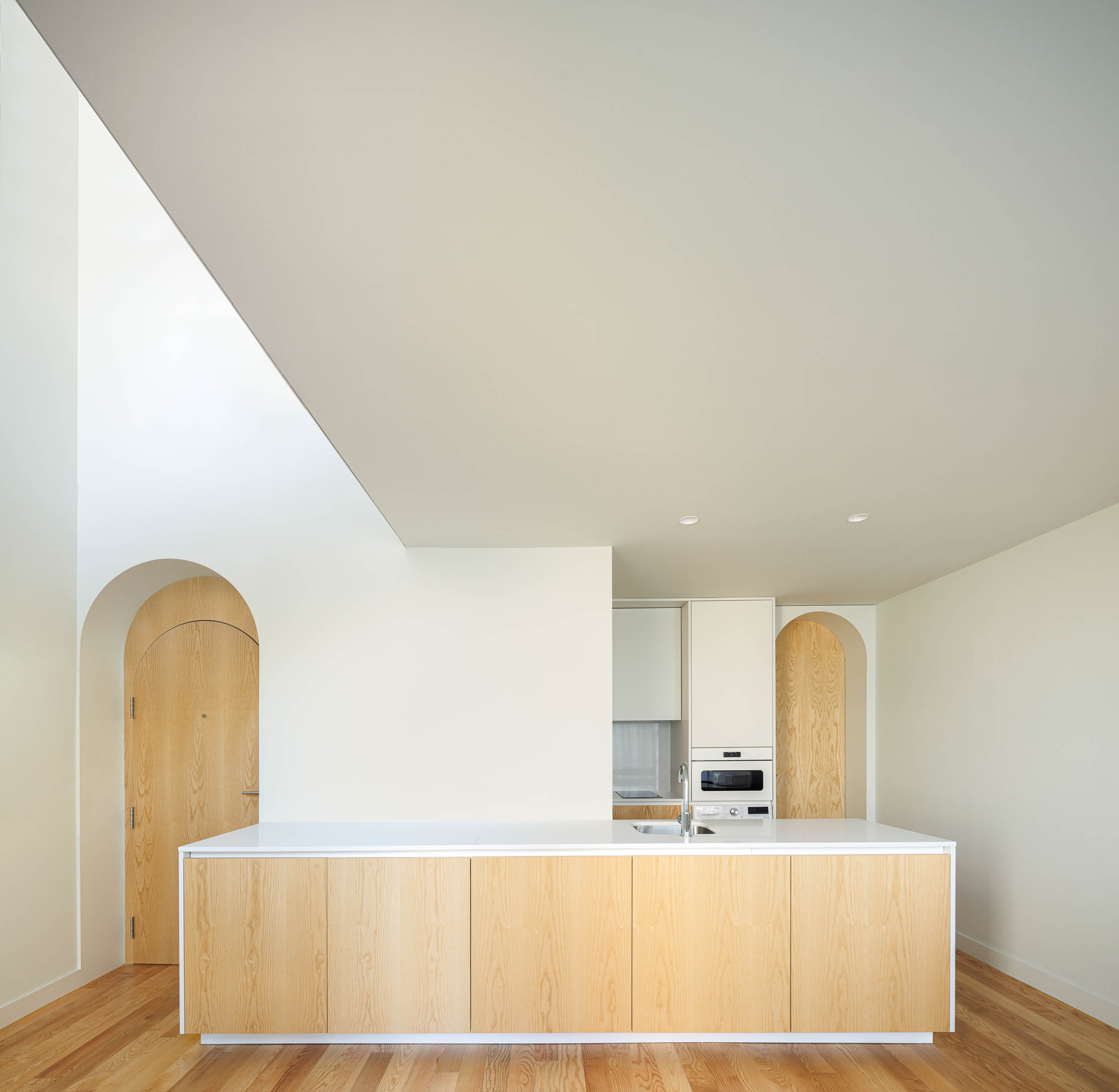 Cozinha com ilha central em madeira clara, porta em arco e claraboia no teto. — Boavista 339 arquitetura Paulo Merlini Architects em Porto
