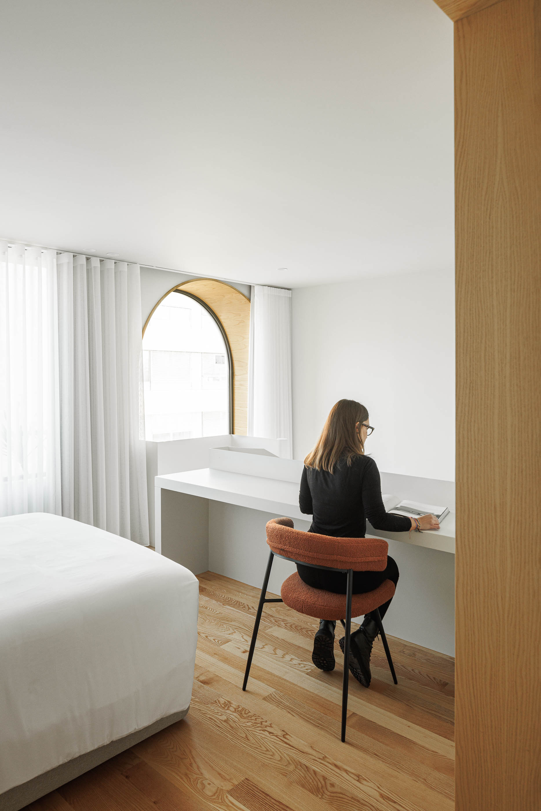 Quarto com secretária branca junto à janela em arco, mulher a ler e cama à esquerda. — Boavista 339 arquitetura Paulo Merlini Architects em Porto