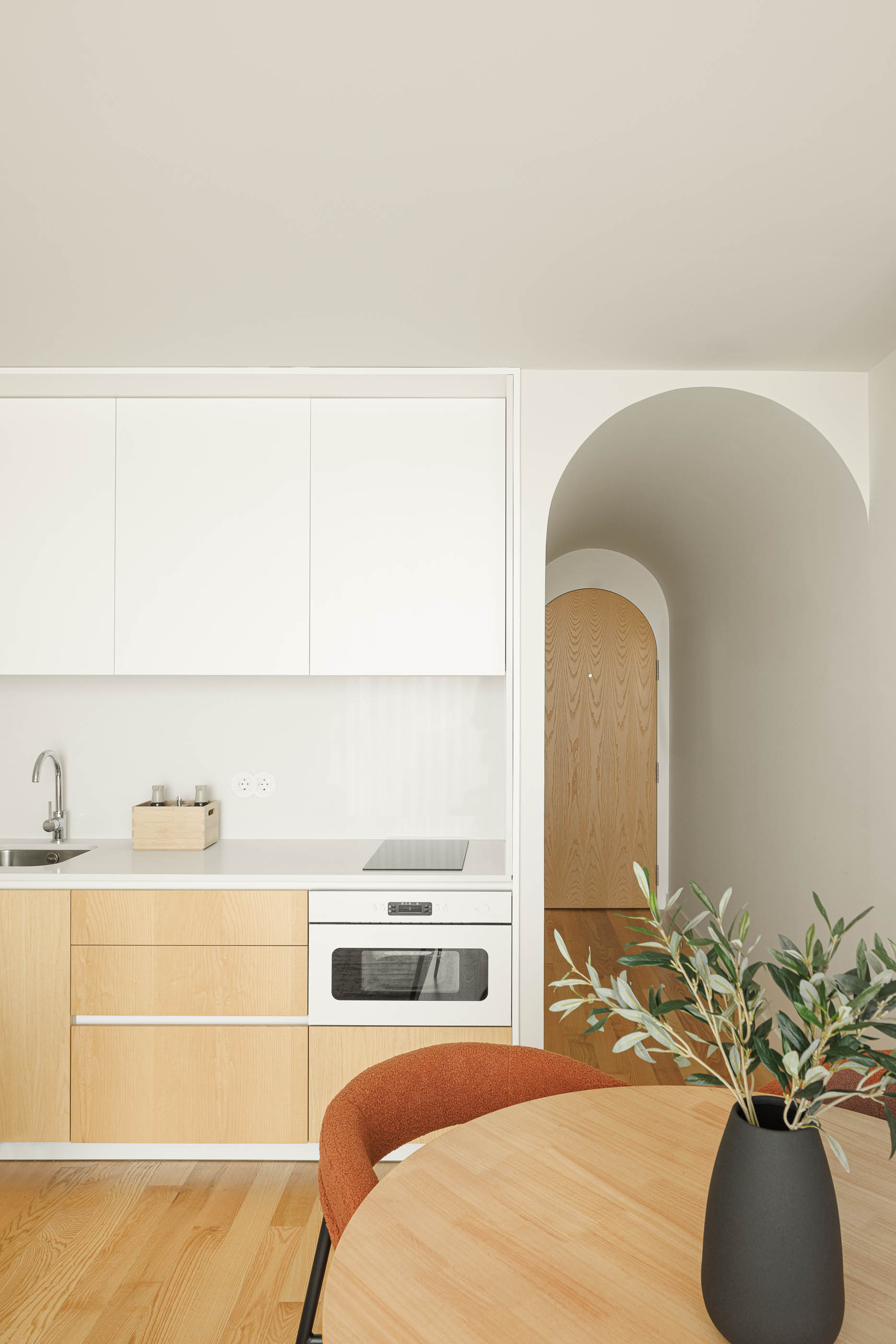 Cozinha com armários brancos e madeira clara, porta em arco e mesa redonda com cadeiras. — Boavista 339 arquitetura Paulo Merlini Architects em Porto