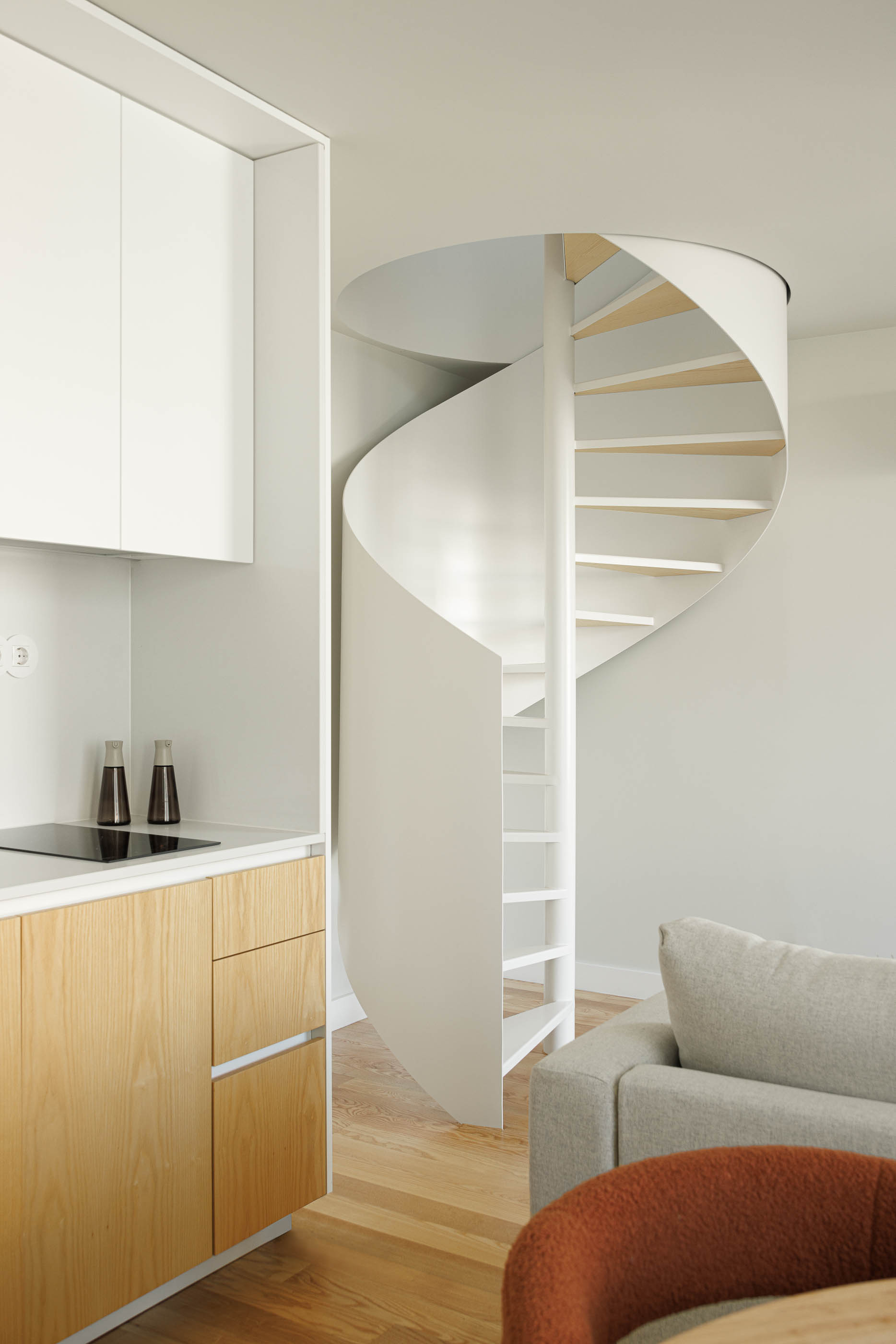 Cozinha e sala de estar com escada em caracol branca, sofá cinzento e armários em madeira clara. — Boavista 339 arquitetura Paulo Merlini Architects em Porto
