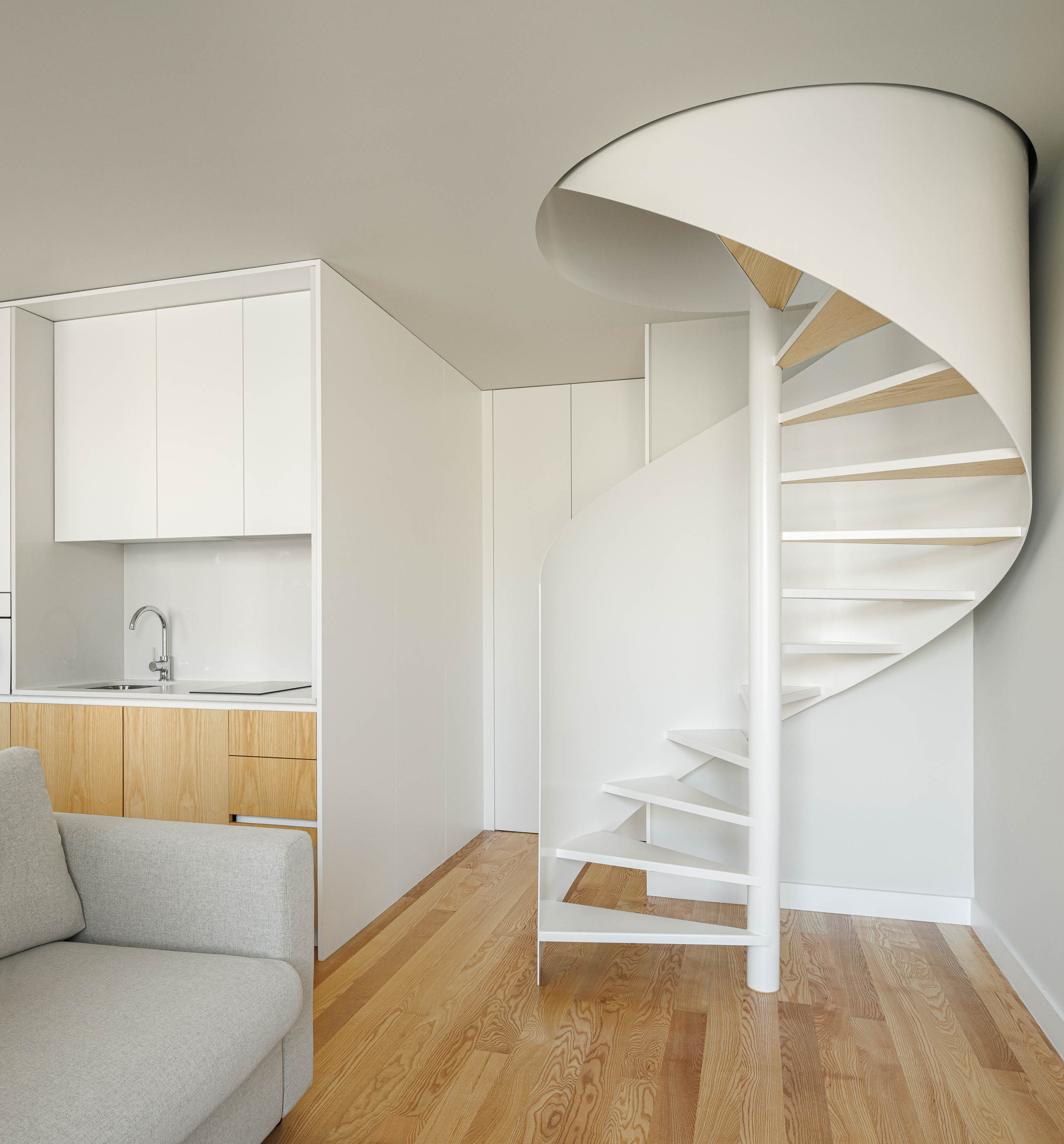 Escada em caracol branca, cozinha com armários brancos e madeira clara, e sofá cinzento. — Boavista 339 arquitetura Paulo Merlini Architects em Porto