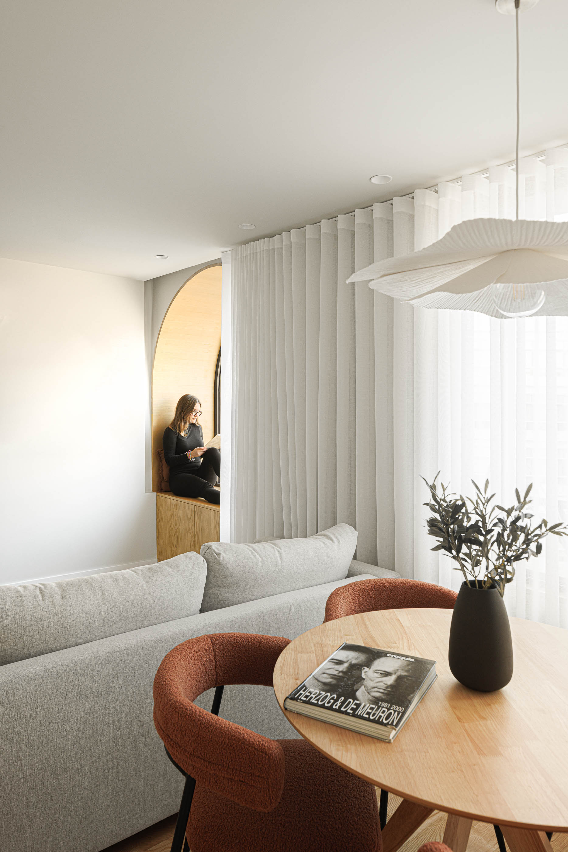 Sala de estar com sofá cinzento, mesa redonda, janela em arco e mulher a ler. — Boavista 339 arquitetura Paulo Merlini Architects em Porto