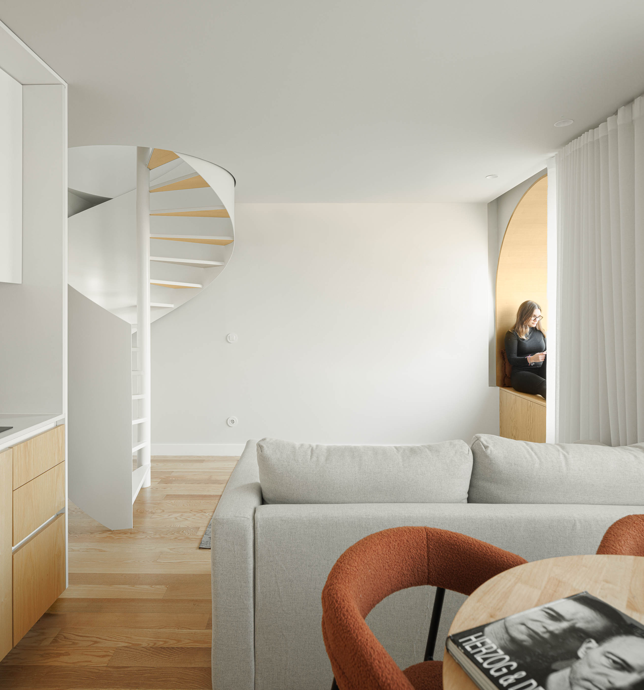 Sala de estar com escada em caracol branca, sofá cinzento, janela em arco e cozinha. — Boavista 339 arquitetura Paulo Merlini Architects em Porto