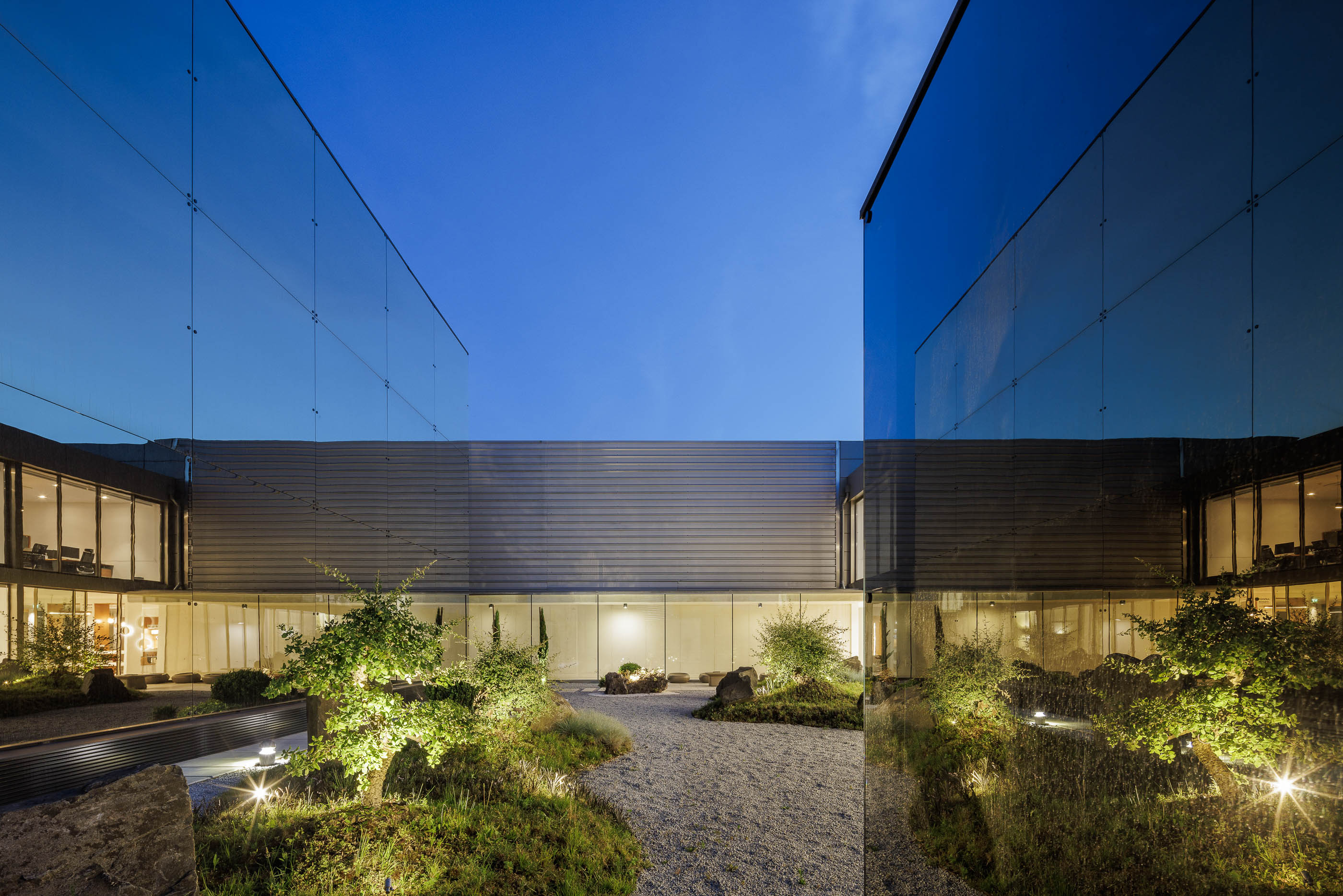 Exterior: Pátio interior com jardim zen e edifícios com fachada espelhada, com iluminação noturna. — Requalificação e Ampliação das Instalações Exporlux arquitetura Espaço Objecto em Águeda