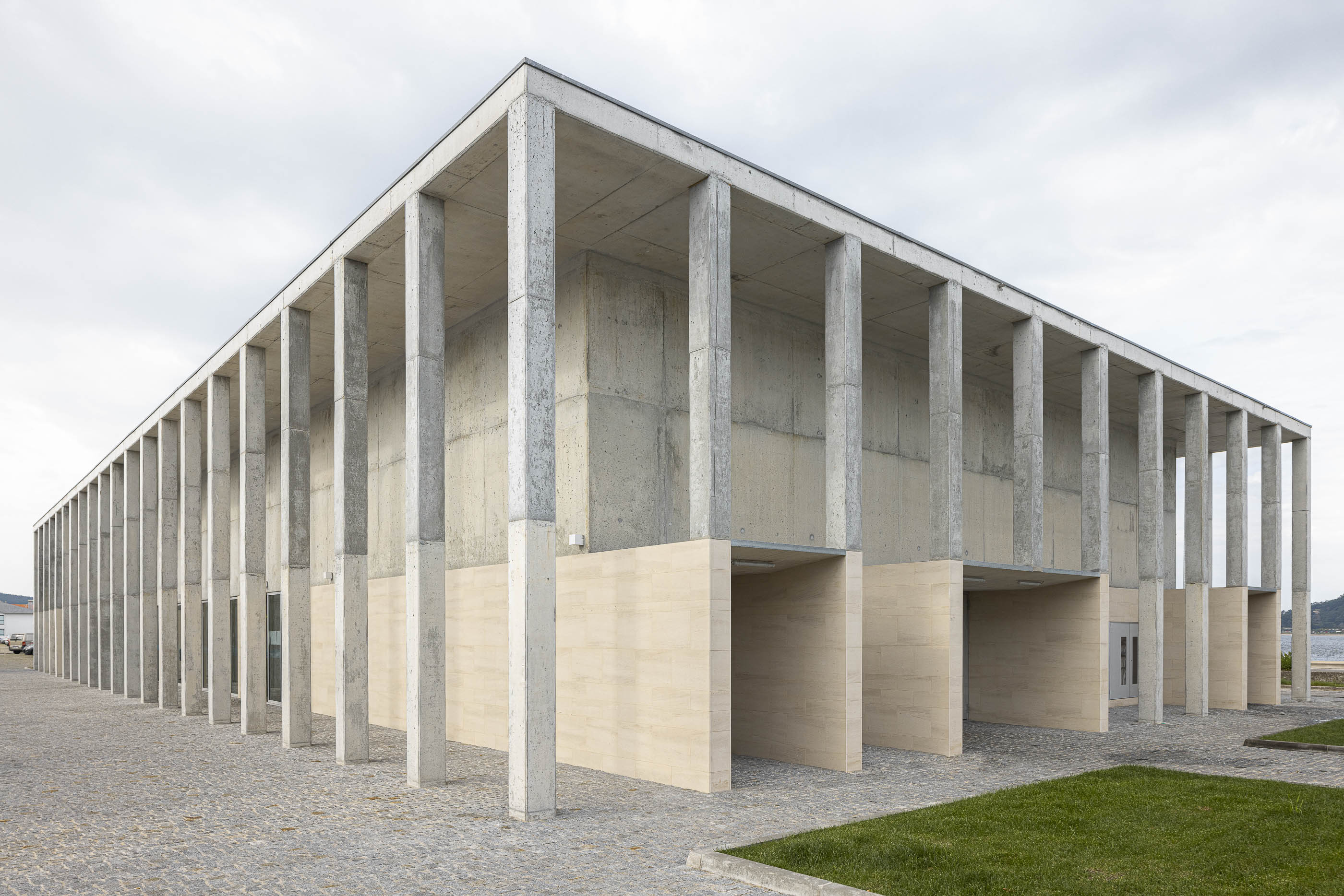 Exterior: Edifício do mercado com estrutura de betão aparente e base em pedra clara, céu nublado. — Mercado Caminha arquitetura Loftspace, Tiago Sousa Arquitecto em Caminha