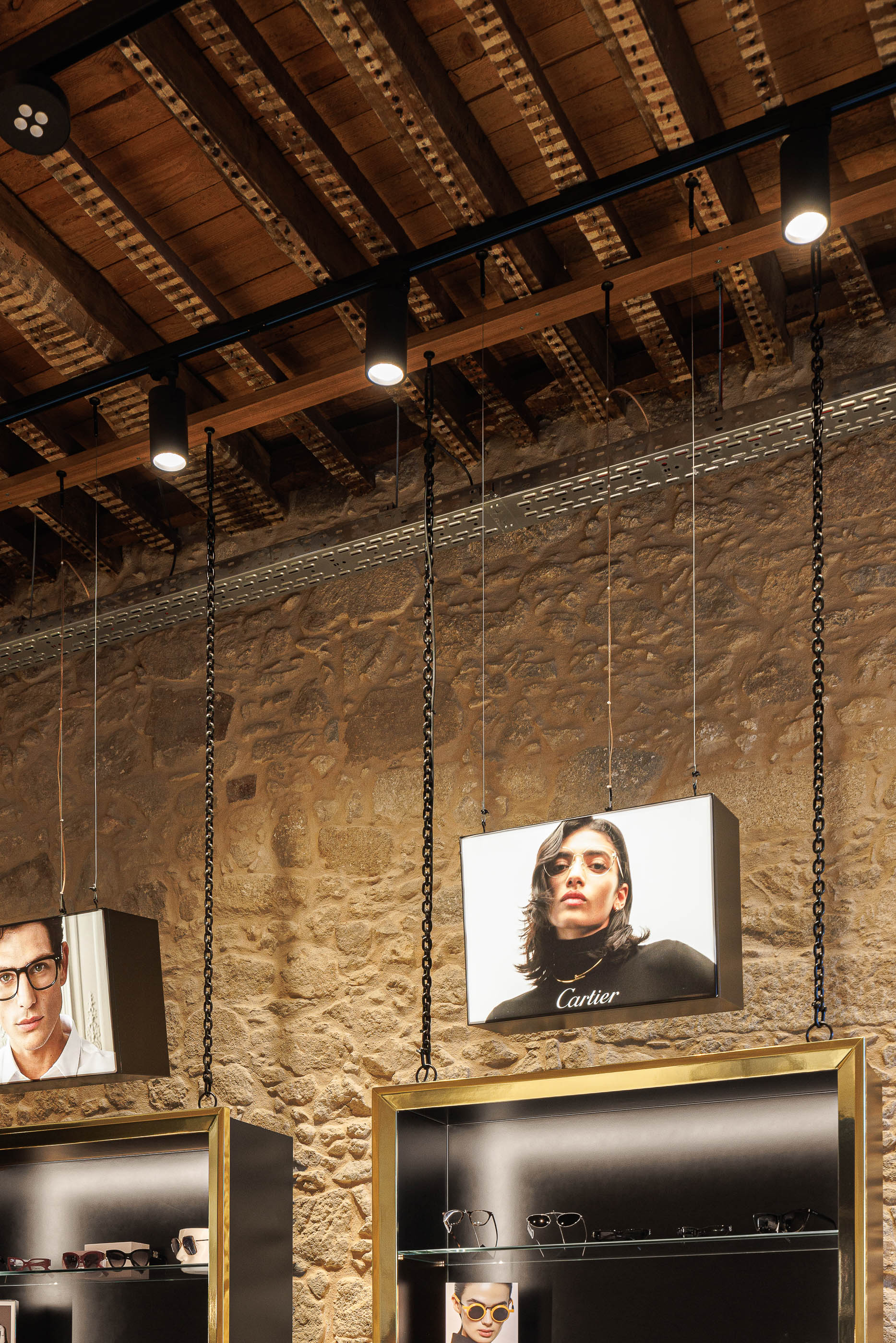 Detalhe: Teto em madeira rústica com vigas, candeeiros cilíndricos pretos e expositores suspensos por correntes. — Jorge Oculista arquitetura Inception Architects Studio em Braga