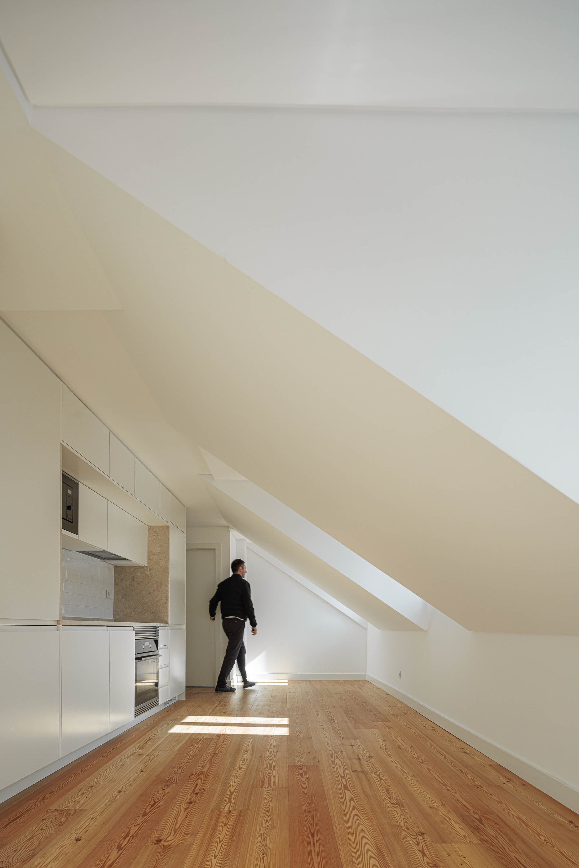 Cozinha: Cozinha em open space com pavimento em madeira clara, armários brancos e pessoa a caminhar. — Anjos 25 arquitetura Pedro Carrilho Arquitectos em Lisboa