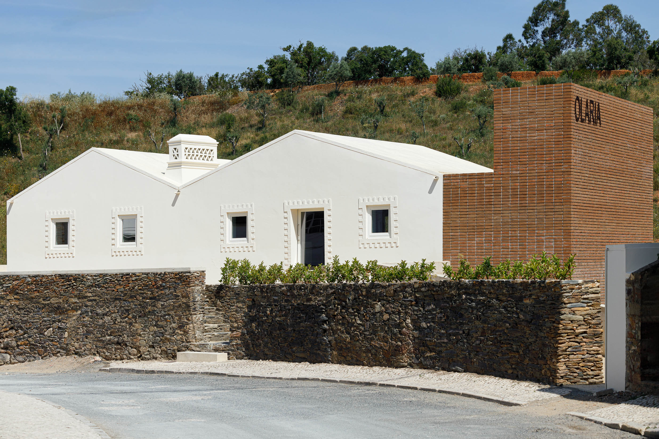 Fachada: Edifício branco de telhado inclinado com detalhes em tijolo e pedra, com a palavra "OLARIA" visível. — Olaria Municipal arquitetura ARDE Arquitetura+Design em Odemira