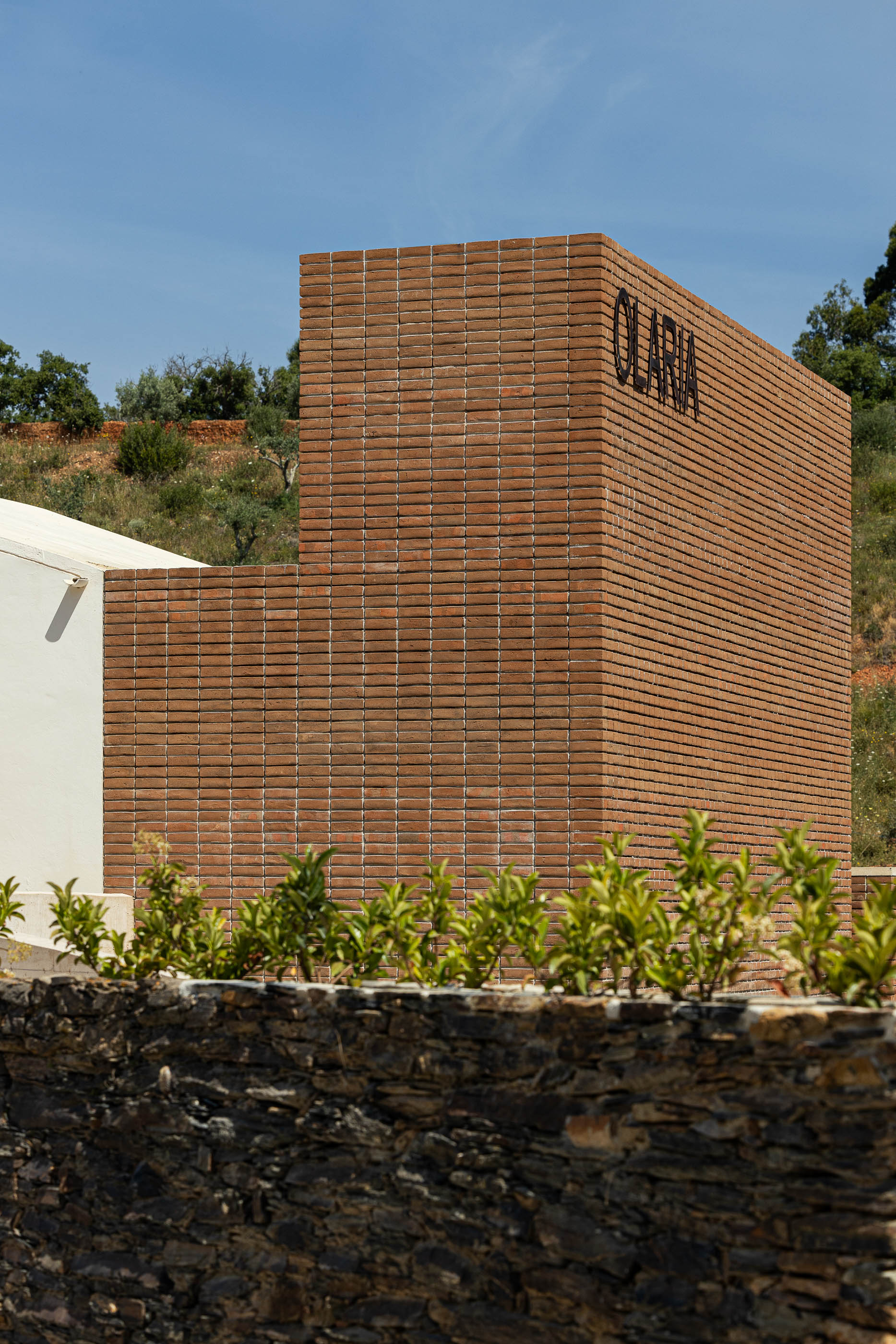 Fachada: Detalhe da parede de tijolo com a inscrição "OLARIA", com vegetação e muro de pedra em primeiro plano. — Olaria Municipal arquitetura ARDE Arquitetura+Design em Odemira