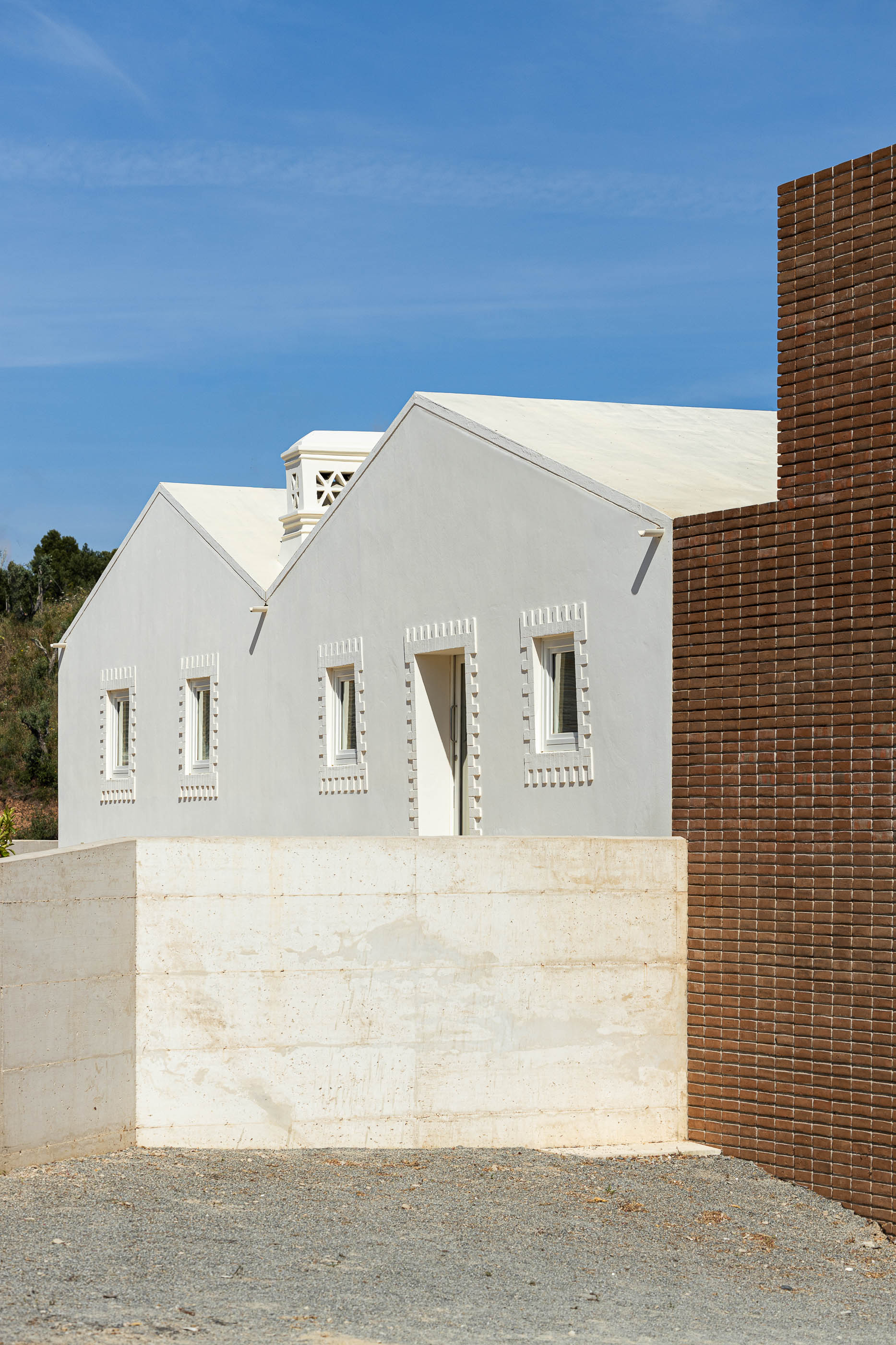 Fachada: Vista lateral do edifício branco com detalhes em tijolo e muro de betão aparente. — Olaria Municipal arquitetura ARDE Arquitetura+Design em Odemira