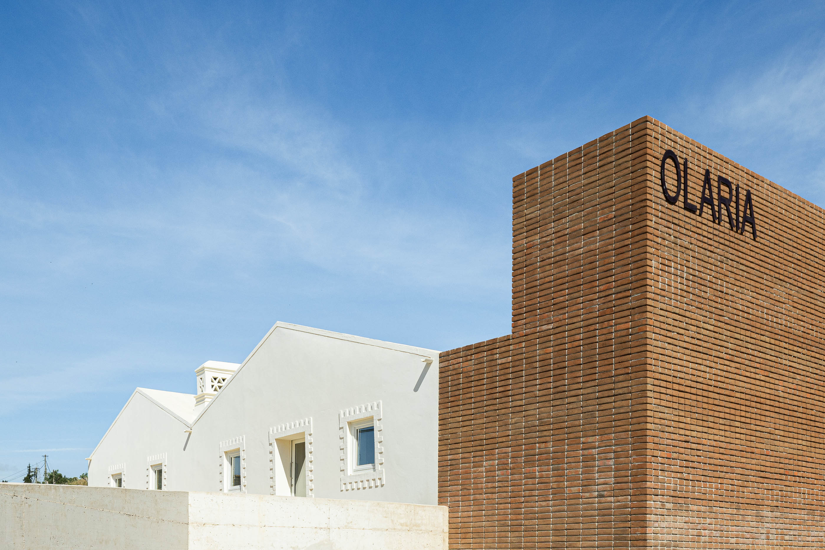 Fachada: Edifício branco com telhado inclinado e torre de tijolo com a palavra "OLARIA" inscrita. — Olaria Municipal arquitetura ARDE Arquitetura+Design em Odemira