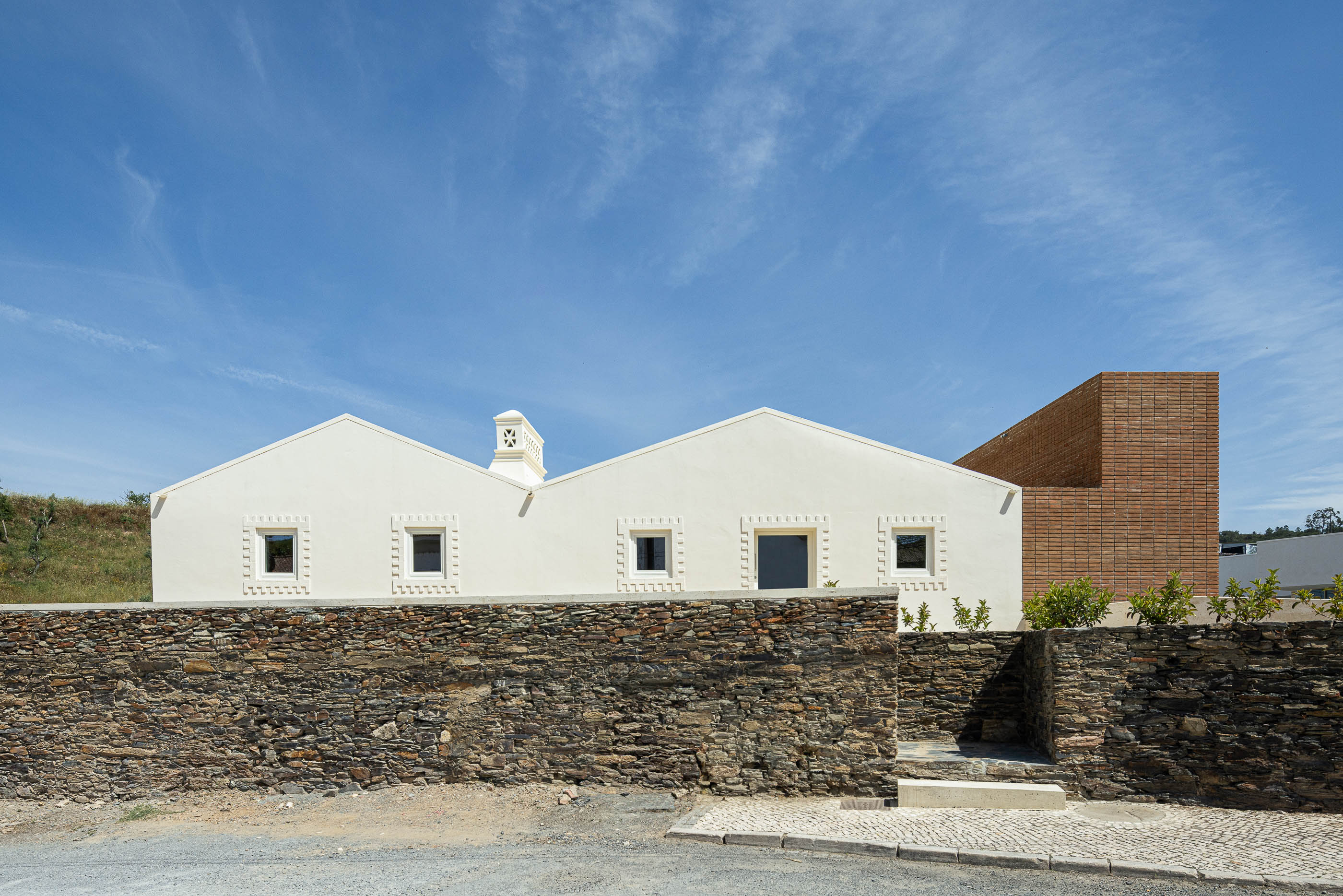 Fachada: Edifício branco com detalhes em tijolo, muro de pedra e vegetação em primeiro plano. — Olaria Municipal arquitetura ARDE Arquitetura+Design em Odemira